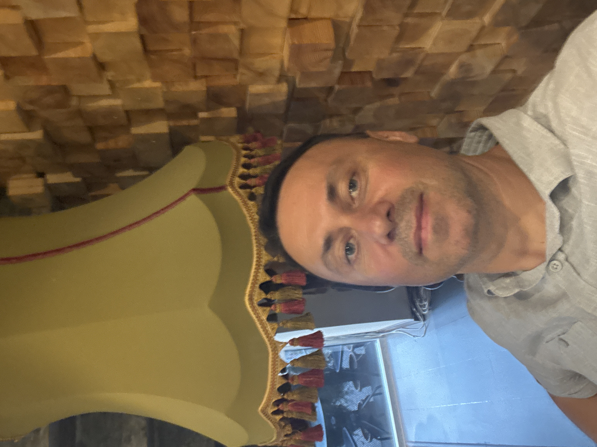 A man is taking a selfie in front of a textured wooden wall and a lamp with tassels. The man is wearing a light-colored collared shirt. The environment appears to be indoors, possibly a restaurant or cafe, with a window in the background that shows reflections of outdoor elements. The wall is made of many small wooden blocks arranged in a repeating pattern. The lamp is large and has a light yellow-green shade with red and gold tassels hanging from its edge. The man is positioned in the foreground, looking at the camera. The lighting suggests it might be evening or an indoor setting with artificial light.