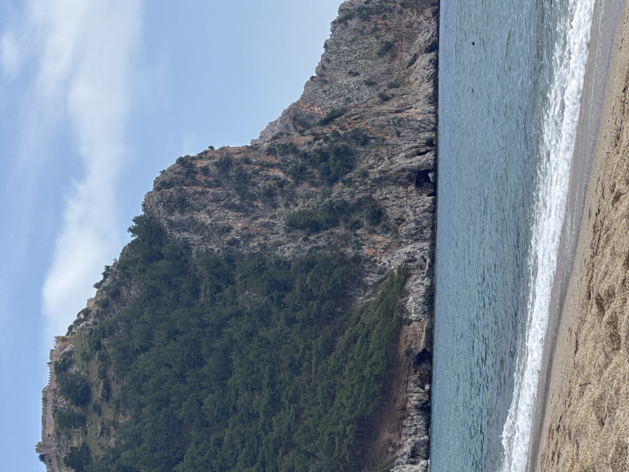 The image depicts a beach scene in Alanya, Turkey. A large, rocky mountain covered in green vegetation dominates the left side of the frame. At the edge of the mountain, closer to the summit, a stone wall, presumably part of Alanya Castle, is visible. The right side of the image shows a sandy beach with waves crashing onto the shore. The water is a clear turquoise color. The sky above is light blue with scattered white clouds. There appears to be a small object (possibly a bird or small boat) in the sea.