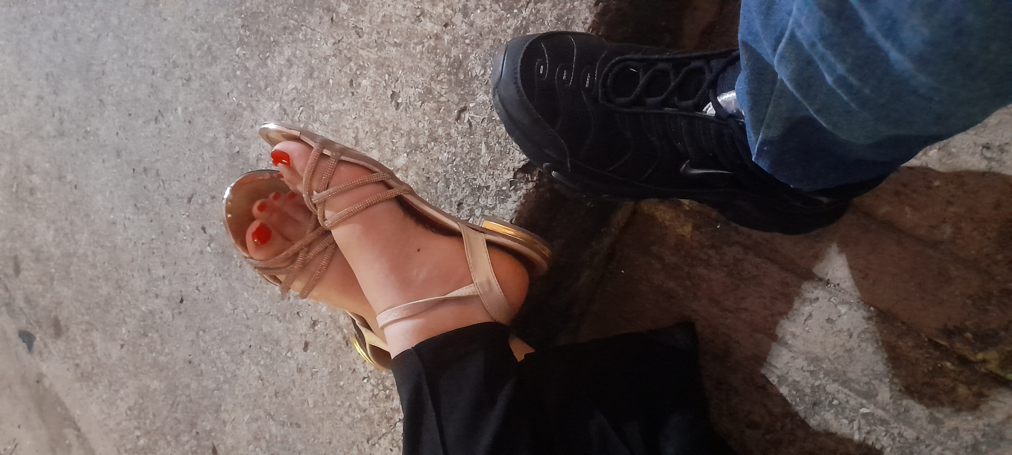 The image captures a close-up, top-down view of two pairs of feet on a textured concrete surface, indicative of an outdoor setting in Juan Griego, Venezuela. On the left, one person wears light gold or champagne-colored strappy sandals with red-painted toenails, paired with black pants. The sandals feature multiple delicate straps across the foot. To the right, another person is seen wearing a black sneaker, identifiable by its distinct rippled design and a visible white Nike swoosh logo on the side, likely a Nike Air Max Plus model. This person is wearing blue jeans. The concrete ground shows variations in tone and texture, with lighter grey areas and darker, possibly shadowed or damp patches. The lighting suggests it is daytime, but with muted sunlight, possibly early morning, late afternoon, or an overcast sky. The image presents a static scene, focusing on the footwear and the ground.