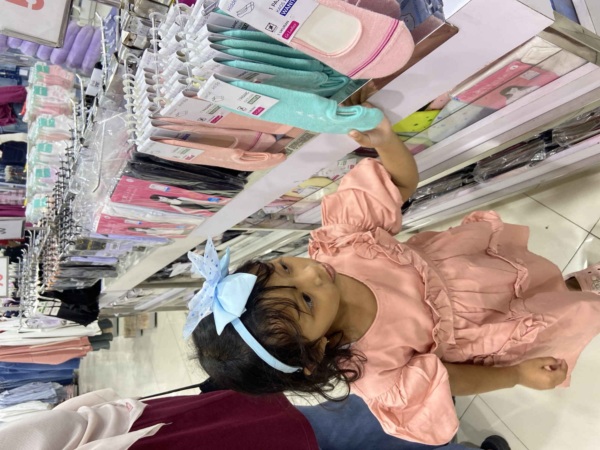 In Sidoarjo, Indonesia, a young girl with curly black hair and a blue polka dot bow headband is browsing socks in a retail store. She is wearing a pink puffy-sleeved dress with ruffles. She reaches out to touch a pair of mint green no-show socks hanging on a display rack. The store is well-lit with rows of clothing and accessories displayed on shelves and hanging racks. Other shoppers are visible in the background, and clothing items are hung on racks in the distance.