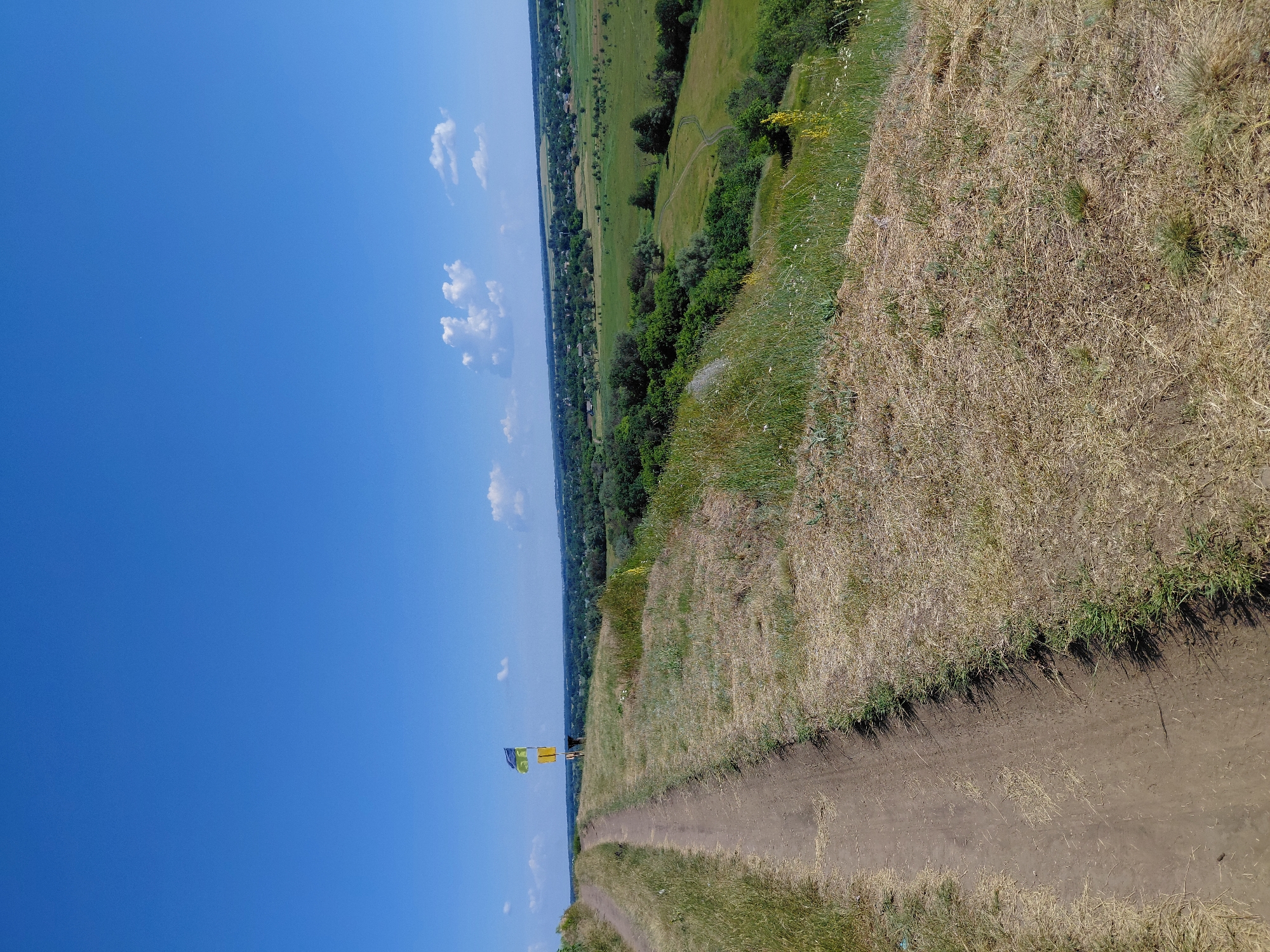 The image depicts a rural landscape under a clear blue sky with scattered white clouds. In the foreground, a dirt path winds its way up a grassy hill, with dry, straw-colored grass dominating the slope. Further up the hill, a flag pole stands with two flags: a blue and yellow flag (likely the Ukrainian flag) and a yellow flag below it. Beyond the hill, a sprawling green valley unfolds, dotted with trees and a village of houses. A few winding paths are visible in the valley floor. The scene suggests a serene, natural environment.