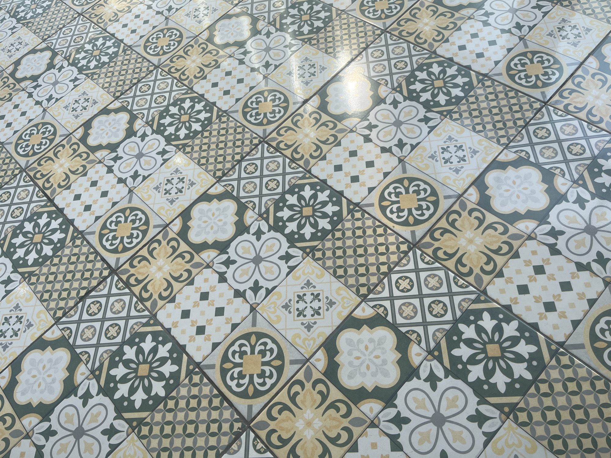 The image shows a close-up, high-angle shot of a floor tiled with a pattern of geometric shapes and floral motifs in shades of green, yellow, white, and gray. The tiles are square and arranged diagonally. The scene is devoid of people and any specific environmental context that would suggest a location in Sidoarjo, Indonesia, or elsewhere. The lighting is uneven, with a bright glare towards the right side of the image, indicating a light source from that direction. The focus is sharp on the tile pattern, with no discernible background.