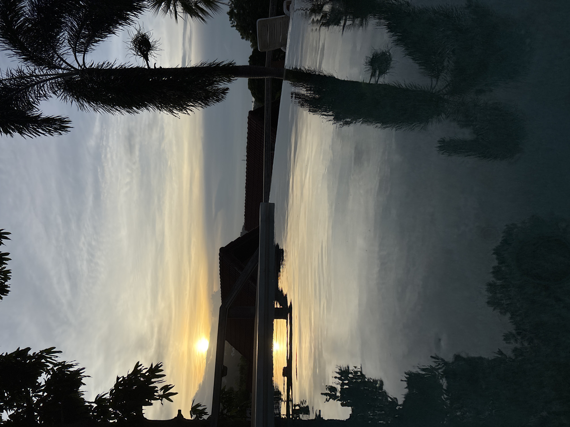 The image depicts a serene sunset scene at an infinity pool, likely located in Ban Bang Rak, Thailand. The sun, a brilliant orb of light, is low on the horizon, casting a warm glow across the sky and reflecting on the pool's surface. Silhouetted against the vibrant sky are the dark shapes of palm trees and foliage, framing the view. In the background, terracotta-tiled roofs of buildings are visible, suggesting a residential or resort setting. The water of the infinity pool creates a mirror-like surface, reflecting the sky, trees, and surrounding environment with remarkable clarity, enhancing the overall tranquility of the scene. There are no people or animals visible in the image. The setting is peaceful and picturesque, capturing the essence of a tropical evening.
