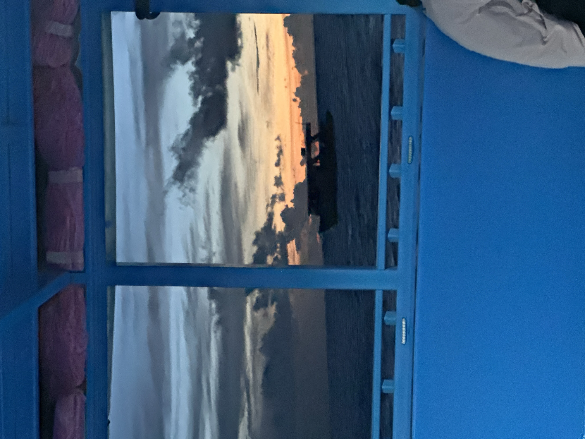 The image captures a view through a blue-framed window of a boat or structure, looking out at a serene ocean scene during sunset. The sky is filled with dramatic clouds, transitioning from shades of gray and deep blue near the top to vibrant oranges and yellows closer to the horizon. A small boat with a dark silhouette is visible on the water, sailing towards the setting sun. The water itself is a deep blue, with gentle waves. In the foreground, the bright blue interior of the boat or structure is visible, with some pink cushions lining the top part of the frame. A person's shoulder and upper arm, wearing a light-colored garment, are partially visible on the right side of the image. The image appears to be taken from within a sheltered area, looking outwards towards the open sea.