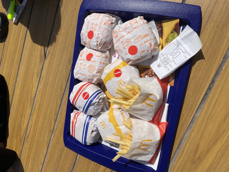 A blue plastic tray sits on a wooden table. The tray contains six wrapped fast food items, two containers of french fries, and a receipt. The fast food items appear to be hamburgers. There are two containers of french fries, one on the left and one on the right of the tray. The receipt is centered in front of the tray, between the containers of french fries. The scene is outdoors and the table is positioned in a place where it receives direct sunlight. This location is near coordinates (latitude: 42.532, longitude: 26.4) in Hadjidimitrovo, Bulgaria.