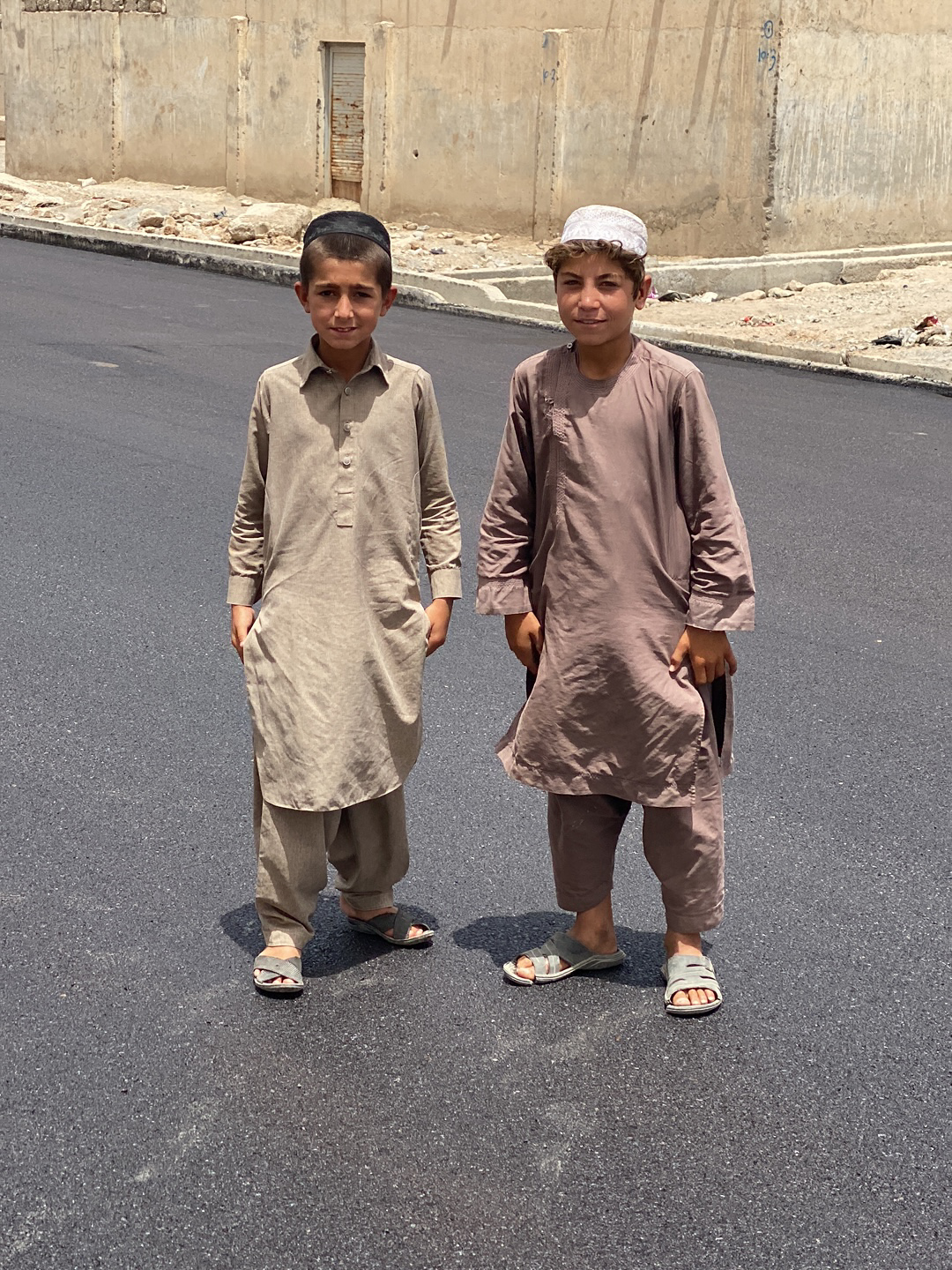 Two young boys are standing on a paved road in what appears to be a city or town setting. The boy on the left wears a beige-colored shalwar kameez and a black skullcap. The boy on the right is dressed in a brown shalwar kameez and a light-colored hat. Both are wearing sandals. The background features a wall or building, and the scene is bathed in bright daylight, suggesting it is outdoors. The exact location is not clear, but the prompt suggests Kandahar, Afghanistan.