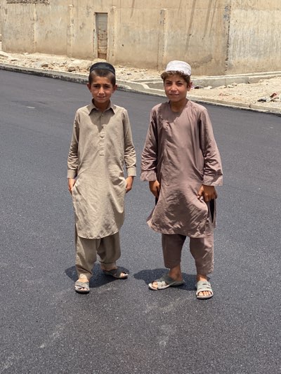 Two young boys are standing on a paved road in what appears to be a city or town setting. The boy on the left wears a beige-colored shalwar kameez and a black skullcap. The boy on the right is dressed in a brown shalwar kameez and a light-colored hat. Both are wearing sandals. The background features a wall or building, and the scene is bathed in bright daylight, suggesting it is outdoors. The exact location is not clear, but the prompt suggests Kandahar, Afghanistan.
