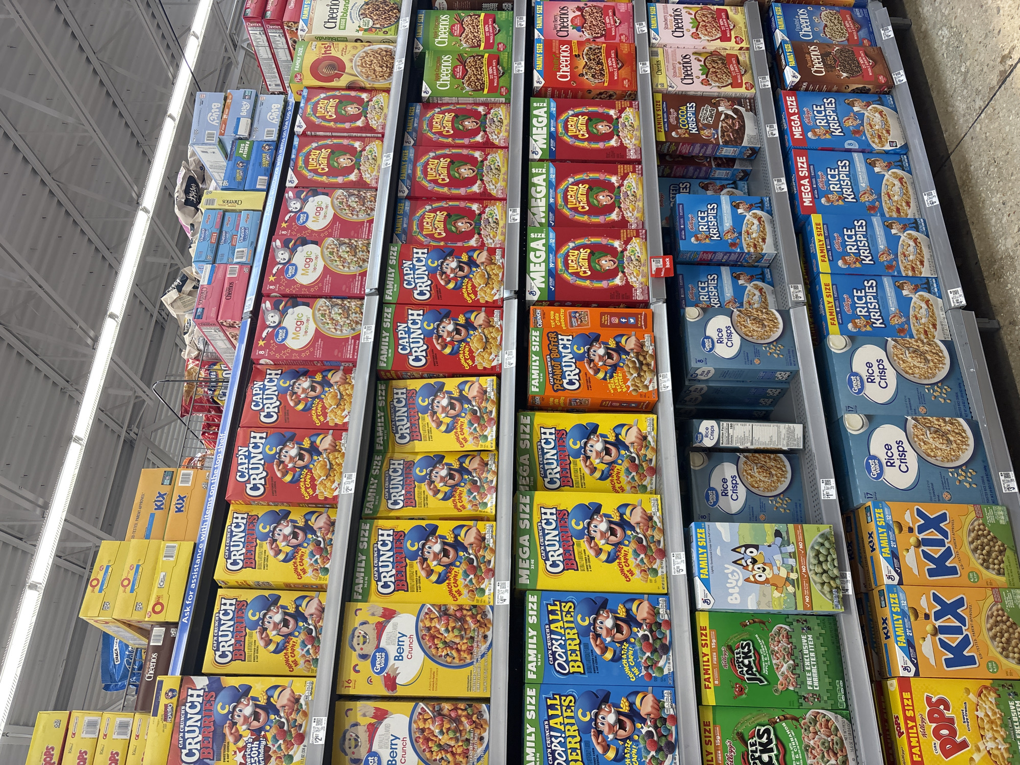 The image shows a well-stocked cereal aisle in a grocery store. The shelves are filled with various brands and sizes of cereal boxes, including Cheerios, Lucky Charms, Cap'n Crunch, Rice Krispies, Kix, and Apple Jacks. The cereals are neatly arranged, with multiple boxes stacked on each shelf. The aisle appears to be well-lit, and the shelves are lined with price tags. While the image does not confirm the city, the store is located in the United States of America as identified by the presence of US-specific cereal brands and packaging. The location in the city of San Antonio, United States of America cannot be confirmed from the image.