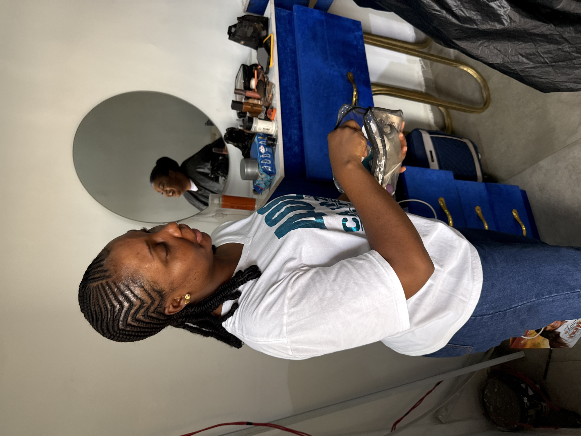 A woman with patterned braids and a gold earring is holding a clear makeup bag in a room with white walls, blue and gold furniture, and a round mirror. She is wearing a white t-shirt and jeans. The mirror reflects another woman in a black top. Makeup and accessories are displayed on a shelf above the woman. Based on the style and context, this is likely in Abuja, Nigeria.