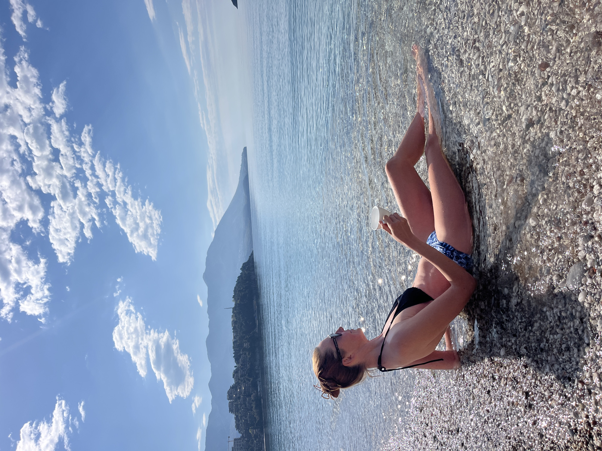 A fair-skinned woman wearing a black bikini top and blue patterned bottoms sits on a pebble beach along the coastline. She is holding a white cup in one hand, tilting her head up, and wearing dark sunglasses. The water laps around her legs, and in the background, there's a view of a mountain range across the water. The sky is a bright blue with some clouds. The location is in Budva, Montenegro.