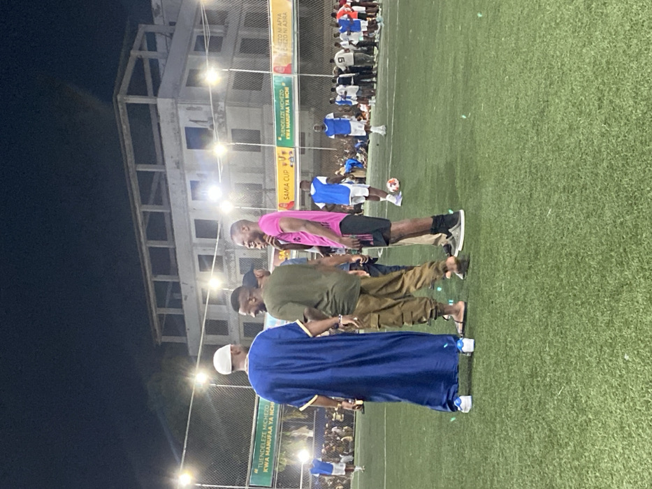 The image shows a group of five men standing on a green artificial turf soccer field at night, located at the coordinates -6.764, 39.258 in Dar es Salaam, Tanzania. The men are interacting and smiling. The man in the blue robe is wearing a white skullcap and is positioned to the left of the frame, while the other four men stand to his right. The man in the pink t-shirt is standing to the far right of the group. The others are wearing casual attire. The man in the green shirt is wearing cargo pants. They are standing in front of a fence with lights overhead. There is a sign in Swahili on the fence and the word "SAMA CUP" is also visible. Several more people are visible in the background, blurred, watching a soccer game on the field. There is a soccer ball on the field to the right. The scene is lit by overhead lighting. The image is taken at night.