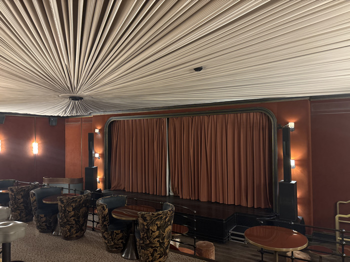 The image depicts an empty, intimate screening room or small theater, identified as the Club de l'Étoile located at 14 Rue Troyon, 75017 Paris, France. The scene features a raised stage positioned at the front, equipped with closed, rust-colored velvet curtains. The stage platform is dark, likely black or a dark wood finish. Flanking the stage are tall, black, columnar structures that incorporate vertical lighting elements, illuminating the adjacent, reddish-brown upholstered walls. Additional wall sconces with two exposed bulbs each provide ambient, warm lighting throughout the room.
The ceiling presents a distinctive design, with light-colored, pleated fabric panels radiating outwards from a central circular fixture, creating a sunburst or fan-like pattern. The seating arrangement consists of multiple dark, ornate armchairs with floral patterns, positioned around small, round, dark wood tables with metallic bases. On the far left, a few lighter-colored, upholstered stools or low chairs with metallic bases are also visible. The floor is covered with a light-colored, speckled carpet. No individuals are present, and no actions or interactions are occurring.