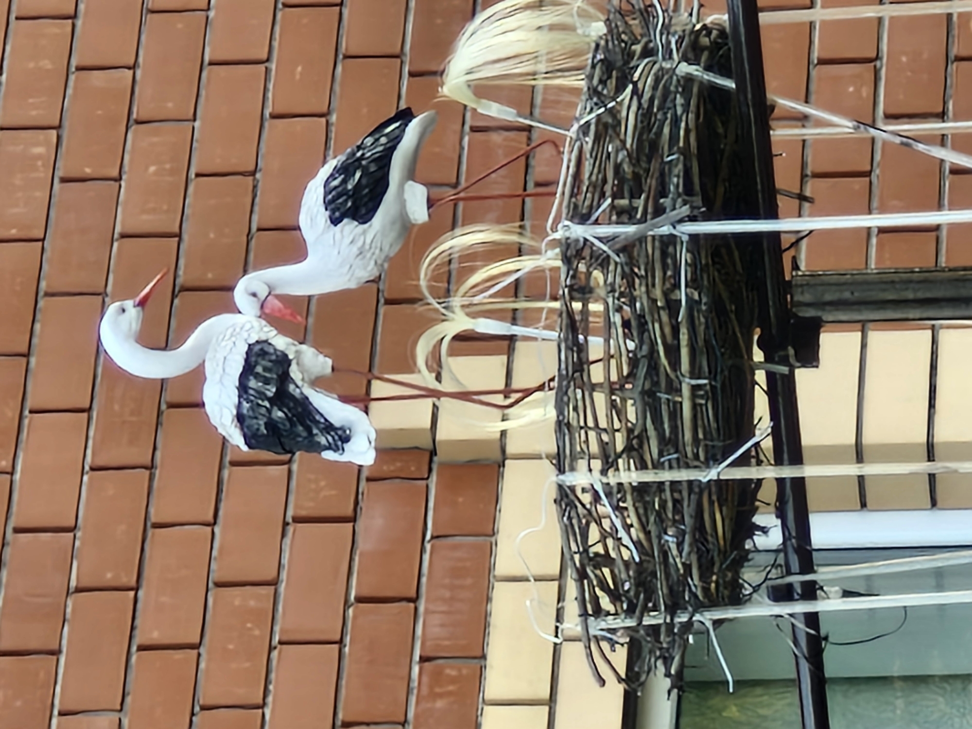 The image shows two decorative white storks with black wing markings and orange beaks standing in a nest made of twigs. The nest is situated in front of a brick wall, with some lighter colored brickwork visible below. The storks appear to be made of plastic or a similar material. The setting suggests an outdoor location, possibly a yard or a rooftop, as indicated by the brick wall and the way the nest is mounted. The overall impression is of a static, decorative display. The location in Buzdyak, Russia, is a detail provided by the user, but not visually inferable from the image itself.