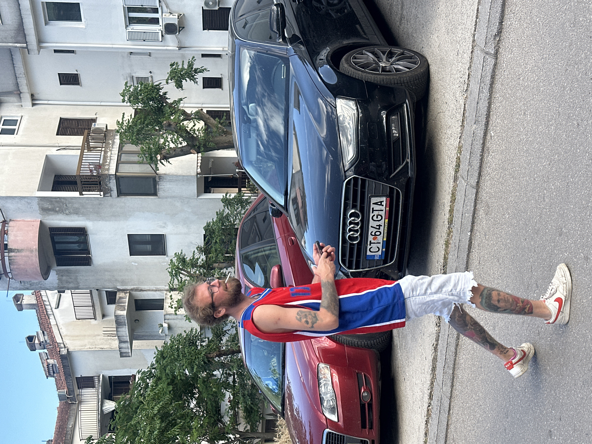 A man stands between two parked Audi cars on a street in Budva, Montenegro. The cars are parked close to a building with multiple windows and balconies. The man, with a beard and glasses, is wearing a red and blue sleeveless jersey, ripped white shorts, and Nike sneakers. He has tattoos on both arms and legs. He is holding a small object in his hands and has a cigarette in his mouth. The license plate of the black Audi shows "CT 64 GTA" and the "RO" country code indicating Romania. The environment appears to be a sunny, urban street.