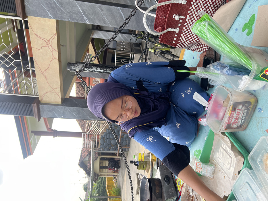 A female individual, wearing a dark blue hijab, light blue long-sleeved shirt with white bow motifs, and matching pants, is seated on a light blue mat decorated with white cactus patterns. She wears light-rimmed glasses and is observed looking down, her right arm extended towards various plastic food containers and utensils. One clear plastic container prominently displays an "Indofood TABURI SAUS BARBEKIU" label. To her left, a portable gas stove with a black pan is positioned on the mat, alongside a yellow bottle of cooking oil and a small gas canister. A red and white woven plastic bag, a black mobile phone, and a brown wallet are also present on the mat near her. In the background, a two-story building with dark grey marbled pillars and beige panels featuring red linear patterns is visible, complete with a balcony and metal railing. A black metal chain fence spans the midground. Beyond the fence, several residential buildings with terracotta-tiled roofs, green bicycles, and distant vegetation are discernible under an overcast sky. This scene is situated in Kecamatan Pati, Indonesia.