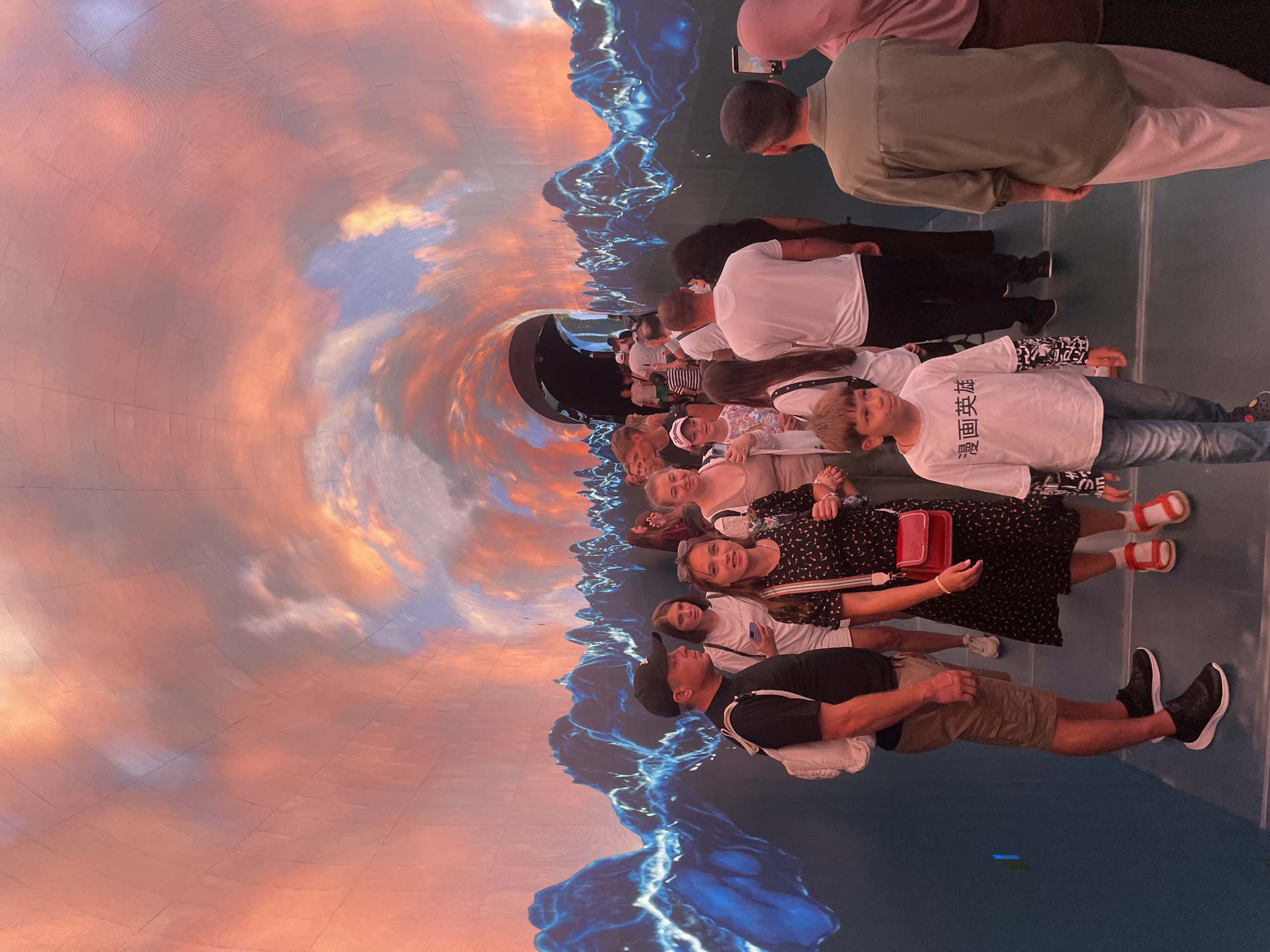 The image depicts a group of people walking through a tunnel-like walkway with illuminated walls. The walls display a vibrant, abstract art piece featuring fiery orange and pink clouds against a backdrop of jagged, blue, mountain-like shapes. The lighting in the tunnel is dominated by these colors, creating an immersive and otherworldly atmosphere.
In the foreground, several individuals are clearly visible, walking towards the viewer and further into the tunnel. A man in a black t-shirt and khaki shorts, wearing a white backpack, is on the left. Next to him, a woman in a black patterned dress with a red purse is smiling. A young boy in a white t-shirt with Japanese characters and jeans is also prominent. Further back, a diverse group of people, including adults and children, are seen walking in the same direction.
The floor of the walkway appears to be a dark, reflective surface, possibly glass or polished material, enhancing the visual impact of the wall art. The overall scene suggests an exhibit or an art installation within a public space.
While the image itself does not provide direct information about its location in Krasnodar, Russia, it is understood that this scene is within the city. The detailed elements observed include the architecture of the walkway, the nature of the digital art display, and the clothing and demographics of the people present. The image captures a moment of leisurely exploration and visual engagement within a technologically enhanced environment.