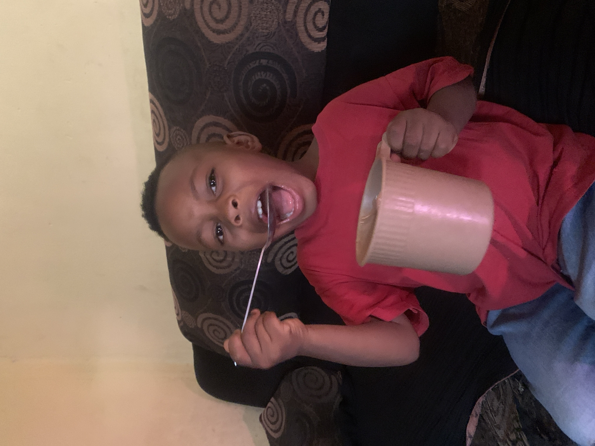 The image captures a candid indoor moment in Jos, Nigeria, featuring a young child, likely a boy, of African descent. The child is positioned on what appears to be a dark-patterned sofa or armchair, wearing a bright red long-sleeved shirt and blue jeans.
The main activity shows the child with a wide-open mouth, revealing his tongue and teeth, in an expressive, almost playful grimace. In his left hand, he holds a silver spoon or fork, pointing towards his mouth as if about to eat. His right hand grasps a large, light-brown or beige mug, suggesting he is either scooping food from it or about to drink. The child's eyes are wide and look directly towards the camera, conveying a lively and engaged emotion.
The background consists of a plain light-colored wall on the left and the dark, swirl-patterned fabric of the furniture on the right. The lighting is soft and diffuse, typical of an indoor setting, and does not indicate a specific time of day. There is no visible text in the image. The scene exudes a spontaneous and joyful energy, capturing a child's playful interaction during mealtime or a snack.