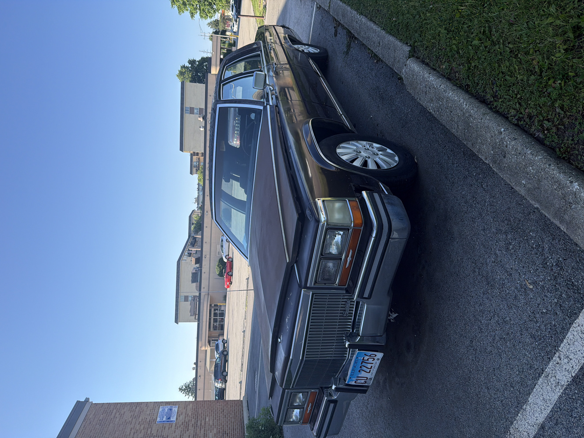 The image shows a dark-colored Cadillac parked on an asphalt surface next to a concrete curb. The car has Illinois license plate number CU 22756. In the background, buildings are visible, and a clear blue sky is overhead. The location appears to be in a parking area. The address is not provided, but the license plate indicates the location is in Illinois, which would include the city of Morton Grove, United States of America.