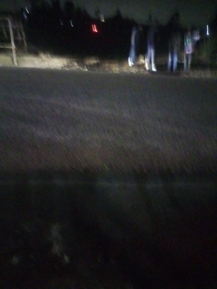 A blurry, dark image shows a road or path with a group of indistinct figures standing on the right side. A structure or object is visible on the left side. Streaks of light appear in the background. The overall scene is difficult to discern due to the lack of focus and low lighting conditions.
