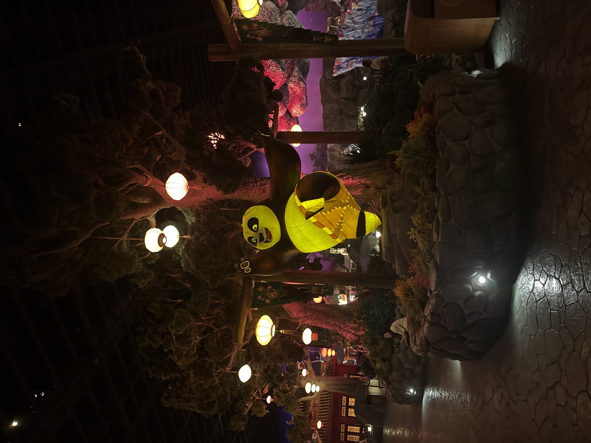 An outdoor nighttime scene depicts a large, illuminated statue of Po, the panda from Kung Fu Panda, striking a martial arts pose. Po is yellow and green, with a prominent belly. He stands on a circular stone pedestal surrounded by lush greenery. Behind him, a large, gnarled tree is adorned with numerous paper lanterns of various sizes and colors, including white, yellow, and red. Some lanterns are lit, casting a warm glow. The background features a stylized, mountainous landscape with purple and pink hues, illuminated by more lanterns and possibly artificial lighting. To the left of the tree, traditional Chinese architecture, including red buildings with ornate windows, is visible. A person is seated on the stone pedestal to the left of Po, looking at their phone. The ground is paved with large, irregular stones. The overall atmosphere is festive and illuminated, suggesting a themed area or attraction within a city like Tongzhou District, China, known for its entertainment venues.