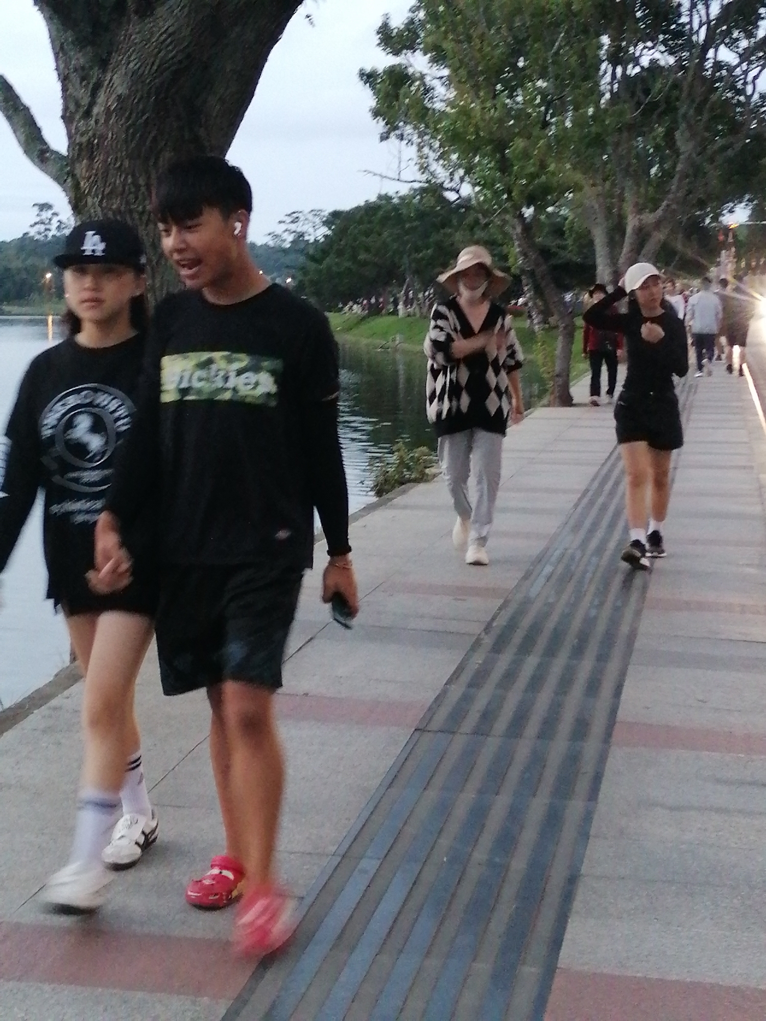 The image depicts a scene along a paved walkway beside a body of water, likely Xuan Huong Lake in Da Lat, Vietnam. In the foreground, a young couple is walking. The male, on the right, wears a black t-shirt with a "Dickies" logo and black shorts. He is holding a smartphone in his right hand. The female, on the left, wears a black t-shirt and a black baseball cap. They are holding hands. Further down the walkway, other individuals are visible, including a woman in a black and white patterned sweater and a straw hat, and another woman in a black shirt and white cap. The environment appears to be a park or recreational area with trees and a cloudy sky. The lighting suggests it is late afternoon or early evening.