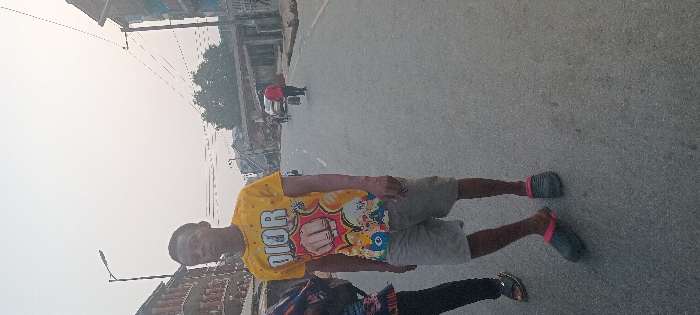 The image displays a street scene in Aba, Nigeria, under diffused daylight. In the foreground, a young male with dark skin and short dark hair stands slightly left of center, facing the camera. He wears a yellow short-sleeved t-shirt with a "DIOR" logo, a cartoon fist, and other colorful characters, along with light grey shorts and dark slip-on clogs featuring pink straps. To his left, the lower body of another person, wearing dark trousers and a dark sandal, is partially visible. The paved road recedes into the distance, occupied by pedestrians and vehicles. A person in a red shirt walks away from the camera in the midground, carrying a dark object. Multi-story buildings with balconies line the left side of the street, while utility poles with overhead wires and a street lamp are visible against a light, hazy sky.