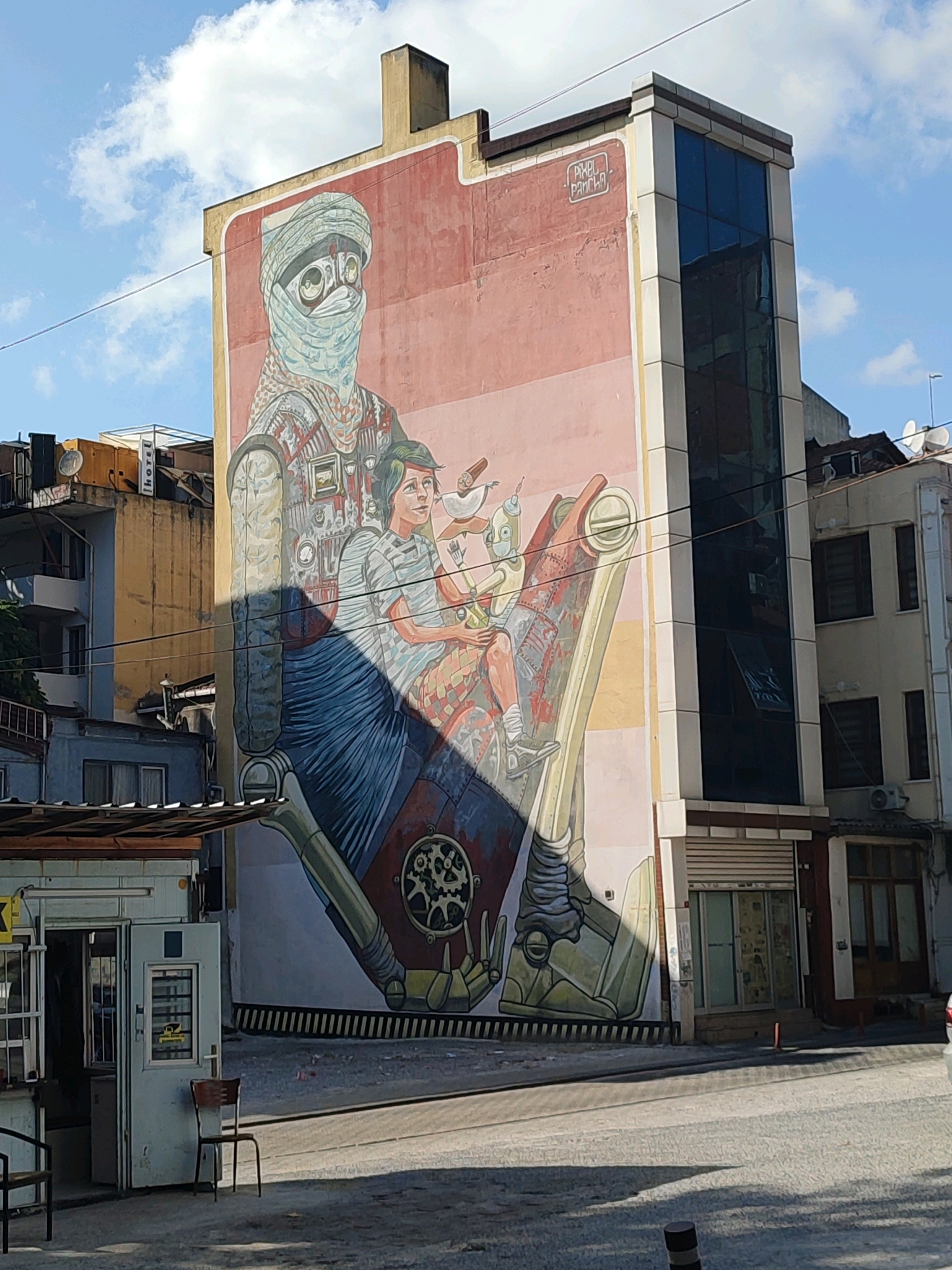 A large mural adorns the side of a building in Kadıköy, Turkey. The mural depicts a robot-like figure wearing a turban and goggles, with a child seated on its lap, holding a tray with drinks. The robot is designed with intricate mechanical details and appears to be made of metal and gears. The child is depicted with a serene expression, looking towards the robot. The mural is painted on a pale pink background with artistic shading and highlights. Above the mural, the building features windows and a sign that reads "PIXEL PANTAN." The surrounding environment shows other buildings, some with visible architectural details like balconies and satellite dishes, and a street with paving stones. The sky above is blue with scattered white clouds, indicating daylight. A small kiosk with an open door and a chair in front of it is visible in the lower left of the frame. The scene suggests an urban setting with a prominent piece of street art.