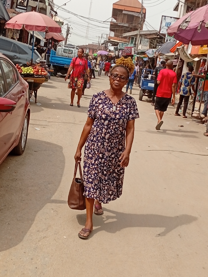 A woman wearing a floral dress and sandals walks toward the camera, holding a brown leather bag. The scene is a busy street, likely a market, with various vendors and pedestrians. There's a red car on the left, and fruit is displayed on a cart nearby. Other people are visible further down the street, some under umbrellas. Buildings and overhead wires fill the background, creating a bustling urban environment. The light suggests a sunny day.