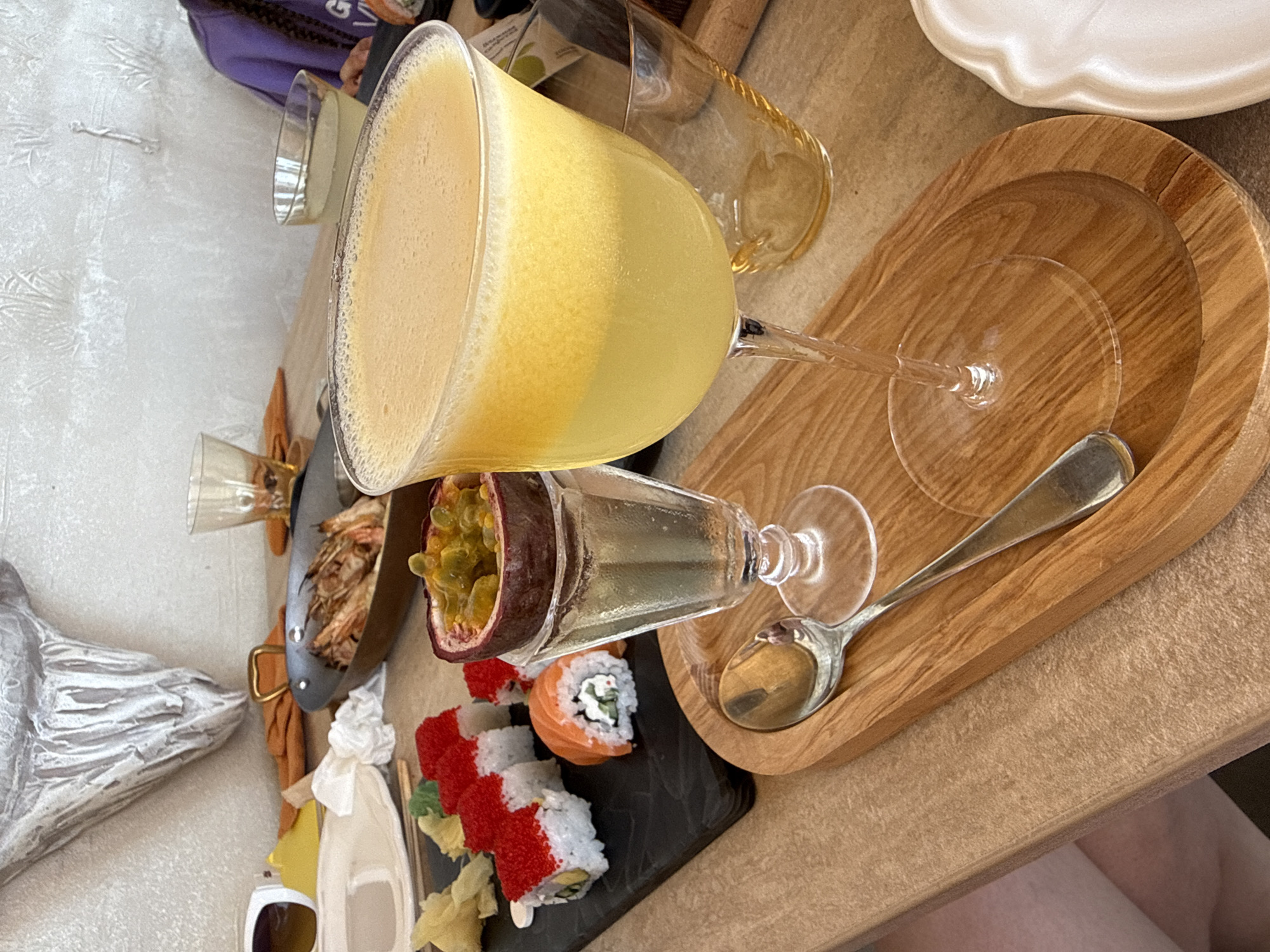 A close-up shot captures a meal on a wooden table, possibly in Alushta, Ukraine. A large, stemmed glass holds a frothy, yellow beverage, likely a cocktail or juice. Next to it, a glass goblet contains a portion of passion fruit, its seeds glistening. A sushi platter with red-capped rolls and a salmon roll is visible on the left, accompanied by ginger. A pan of cooked shrimp sits behind the sushi. In the background, a person wearing a purple garment with braids is partially visible. The table is set with various plates and glasses, suggesting a dining setting.