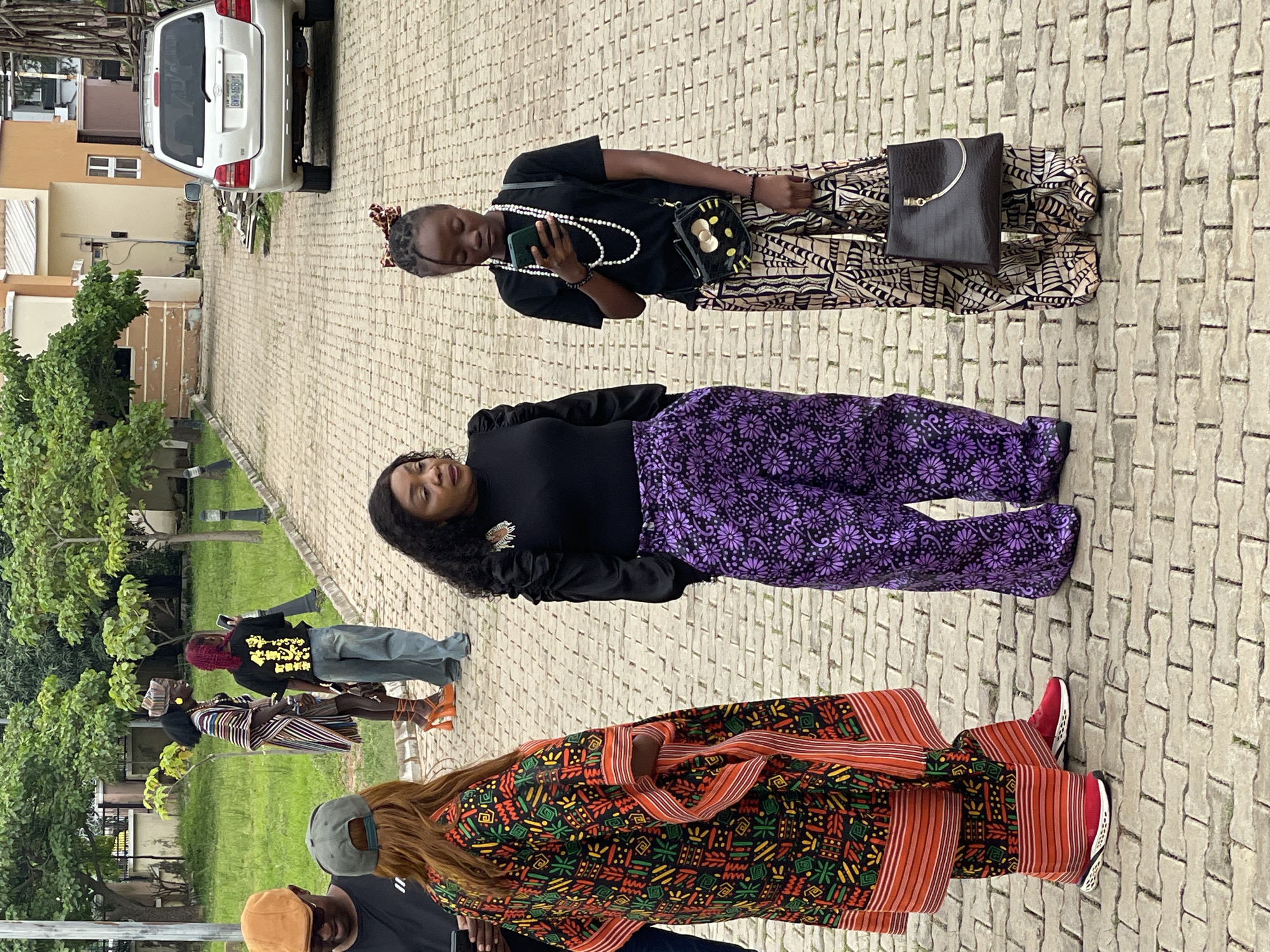 In Barakin Lerawi, Nigeria, three women are standing on a paved outdoor area. The woman on the left is wearing a vibrant, multi-colored patterned kaftan with orange trim and red sneakers. She is looking down at her phone. In the center, a woman is wearing a black top and high-waisted purple pants with a floral pattern. She is looking to her right. To the right, a young woman is wearing a black t-shirt, a long pearl necklace, and patterned pants. She is looking down at her phone and holding a black handbag with a gold chain. In the background, a silver SUV is parked on the right side of the frame. Trees and foliage are visible behind the women and the vehicle. The area appears to be a driveway or courtyard with a cobblestone-like pavement.