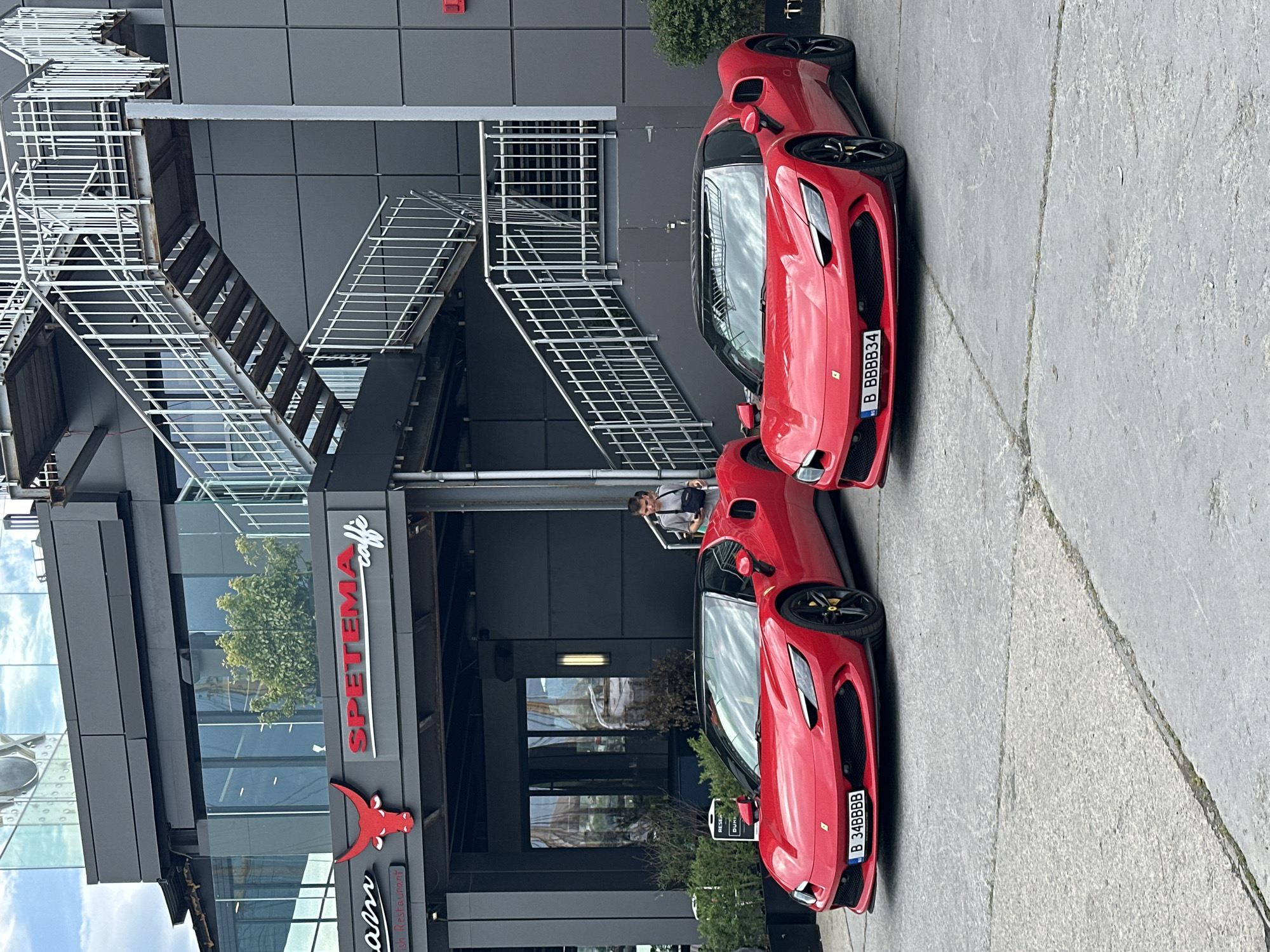 The scene depicts two red Ferrari sports cars parked outside the "SPETEMA caffe" restaurant, situated in Varna, Bulgaria. The building features modern architecture with dark gray panels, glass windows, and a prominent metal staircase on the exterior. The restaurant's logo, featuring a red bull, is visible. A person is standing on the staircase. The cars are parked on a concrete surface. The license plates on the Ferraris are "B 48888" and "B BBBBB34".