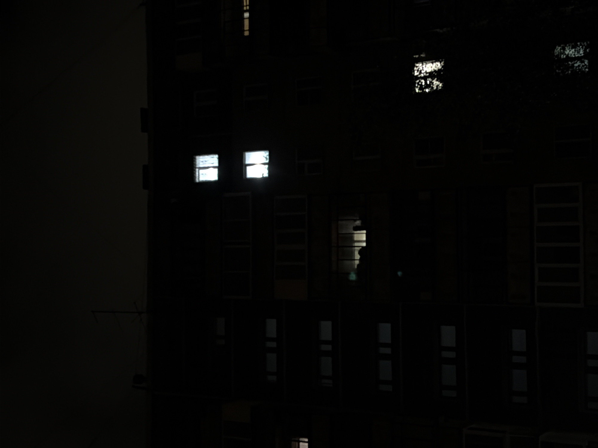 A nighttime exterior shot of a dark, multi-story apartment building shows several illuminated windows against the black facade. The building appears to be made of concrete. At the top of the building, a television antenna is visible. The sky is dark, suggesting it is nighttime. The scene is devoid of people or activity. The image appears to be taken from a low angle, looking up at the building. There are no signs or identifying marks that would indicate its specific location within Chelyabinsk, Russia.