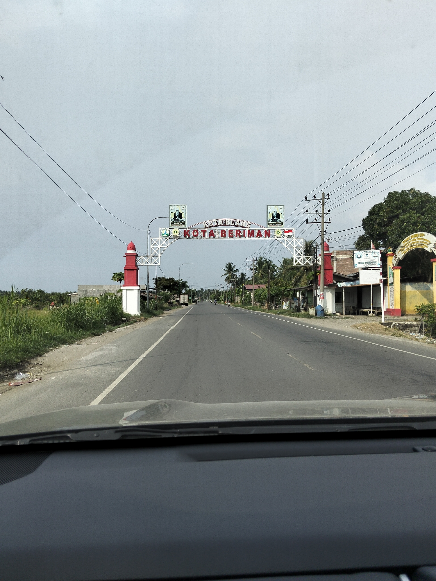The image captures a view from inside a vehicle looking out at a street scene in Uleepusong, Indonesia. In the foreground, the dashboard of the vehicle is visible. The road ahead is paved and appears to be a main thoroughfare with a white line marking the lane. On the left side of the road, tall green grass and some debris are present. On the right side of the road, there are various structures including buildings, a red and white lighthouse-like monument, and an archway with yellow and red accents. A prominent white archway spans the road in the distance, bearing the text "KOTA" and what appears to be "BERIMAN" in Indonesian. Above the archway, there are crests with images and text. Utility poles with numerous wires are scattered along the roadside. The sky is overcast. Several vehicles, including motorcycles and a truck, are present on the road. Palm trees and other greenery are visible along the sides of the road, indicating a tropical or subtropical climate. The overall impression is of a gateway to a town or city.