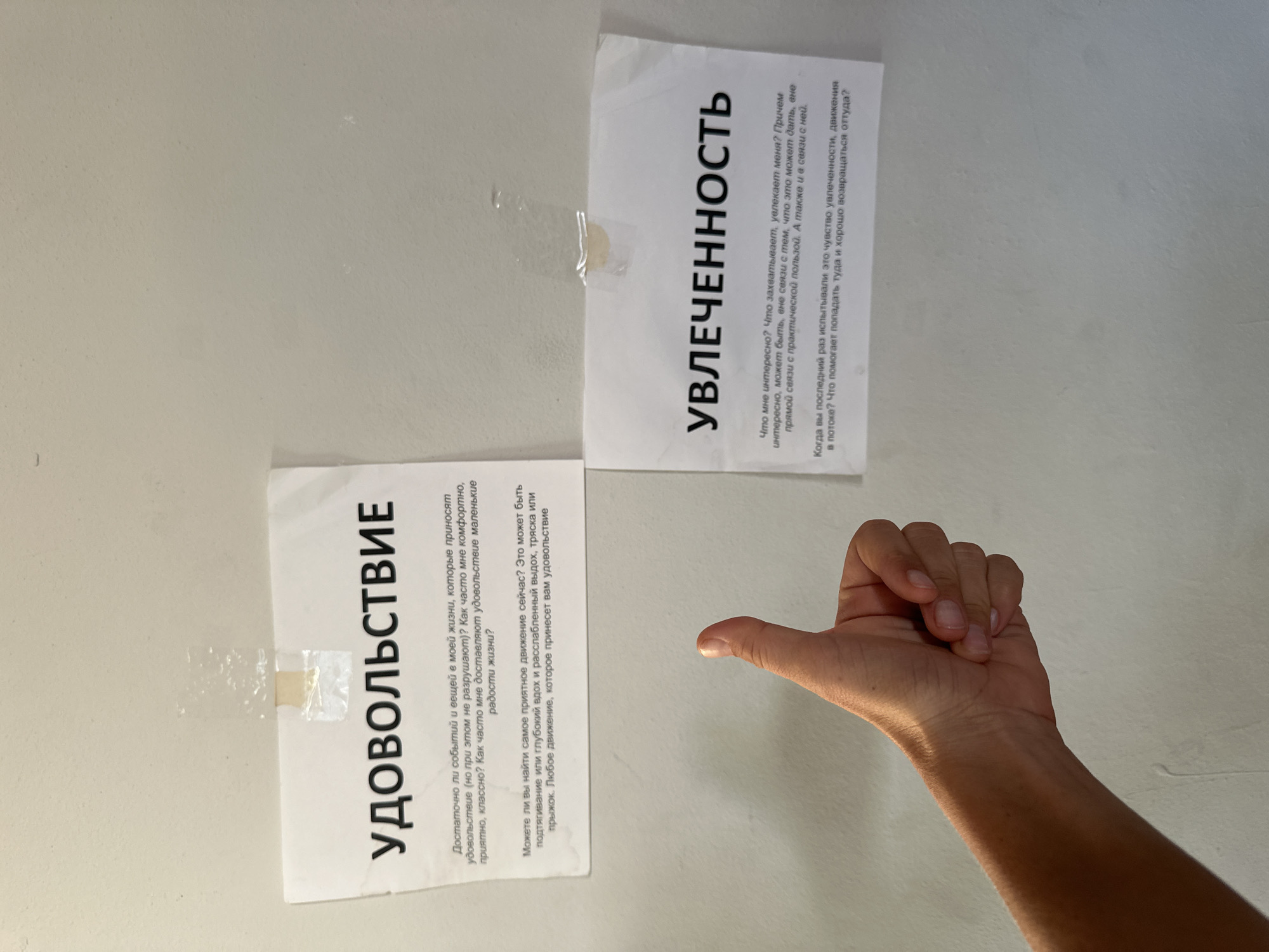 A human arm and hand is visible in the foreground of this image, with the hand in a thumbs-up gesture. The arm and hand are positioned in the lower right quadrant of the frame, casting a shadow on the light-colored wall behind it. Two sheets of paper are affixed to the wall with tape. The paper on the left is titled "УДОВОЛЬСТВИЕ" (Pleasure) and contains text in Russian asking questions about enjoyment and life's small joys. The paper on the right is titled "УВЛЕЧЕННОСТЬ" (Engagement/Enthusiasm) and also contains Russian text, posing questions about interests and the feeling of being in a flow state. The lighting suggests an indoor setting, possibly during the day. The overall impression is one of contemplation or self-reflection, prompted by the questions on the posters.