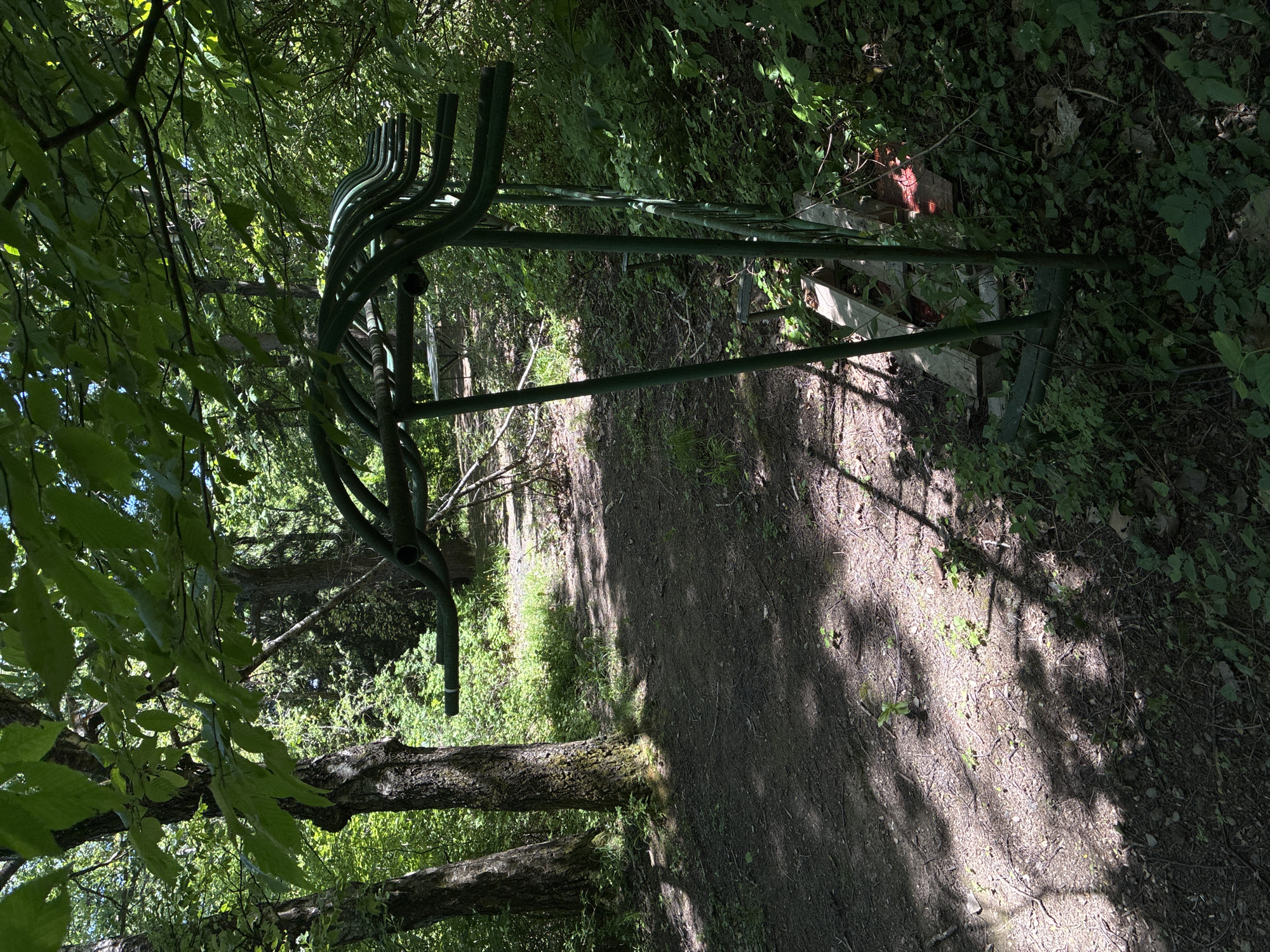 A dirt path, partially shaded by trees, stretches into the distance. A green metal structure, potentially a bike rack, stands to the side of the path, partially obscured by vegetation. The location appears to be a park or wooded area, likely in Sofia, Bulgaria. The image shows no people or animals. There are no signs of any interactions or activities.