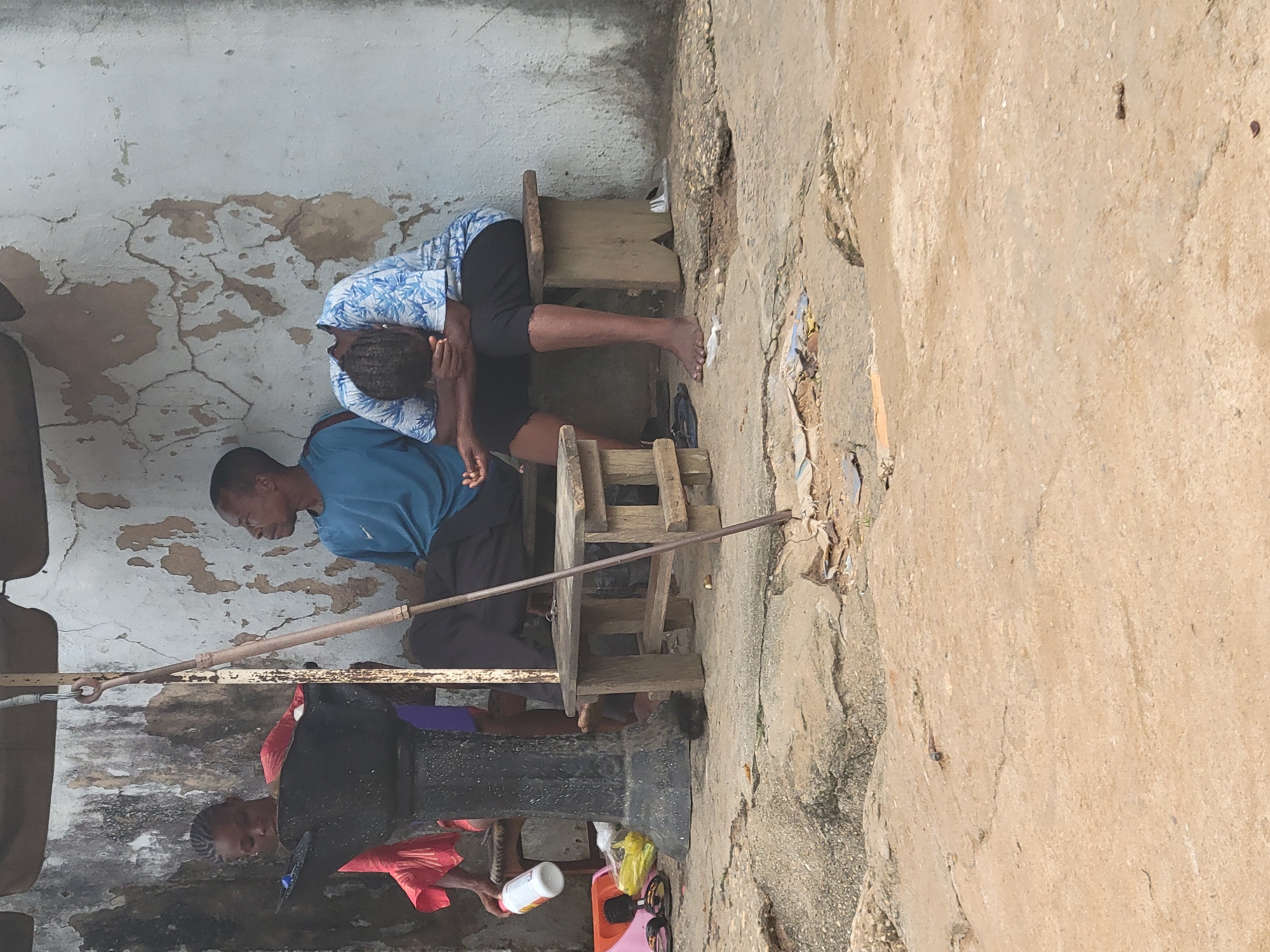 The image shows two individuals, one man and one woman, seated outside a building with a distressed, cracked white wall in Aba, Nigeria. The man is wearing a blue shirt and black pants, while the woman is wearing a blue and white patterned shirt and black pants. Both are seated on small wooden stools. An old water pump with a handle is located nearby. Various objects such as a plastic container and other miscellaneous items are scattered around the base of the pump. The ground appears to be concrete, cracked, and showing signs of wear.