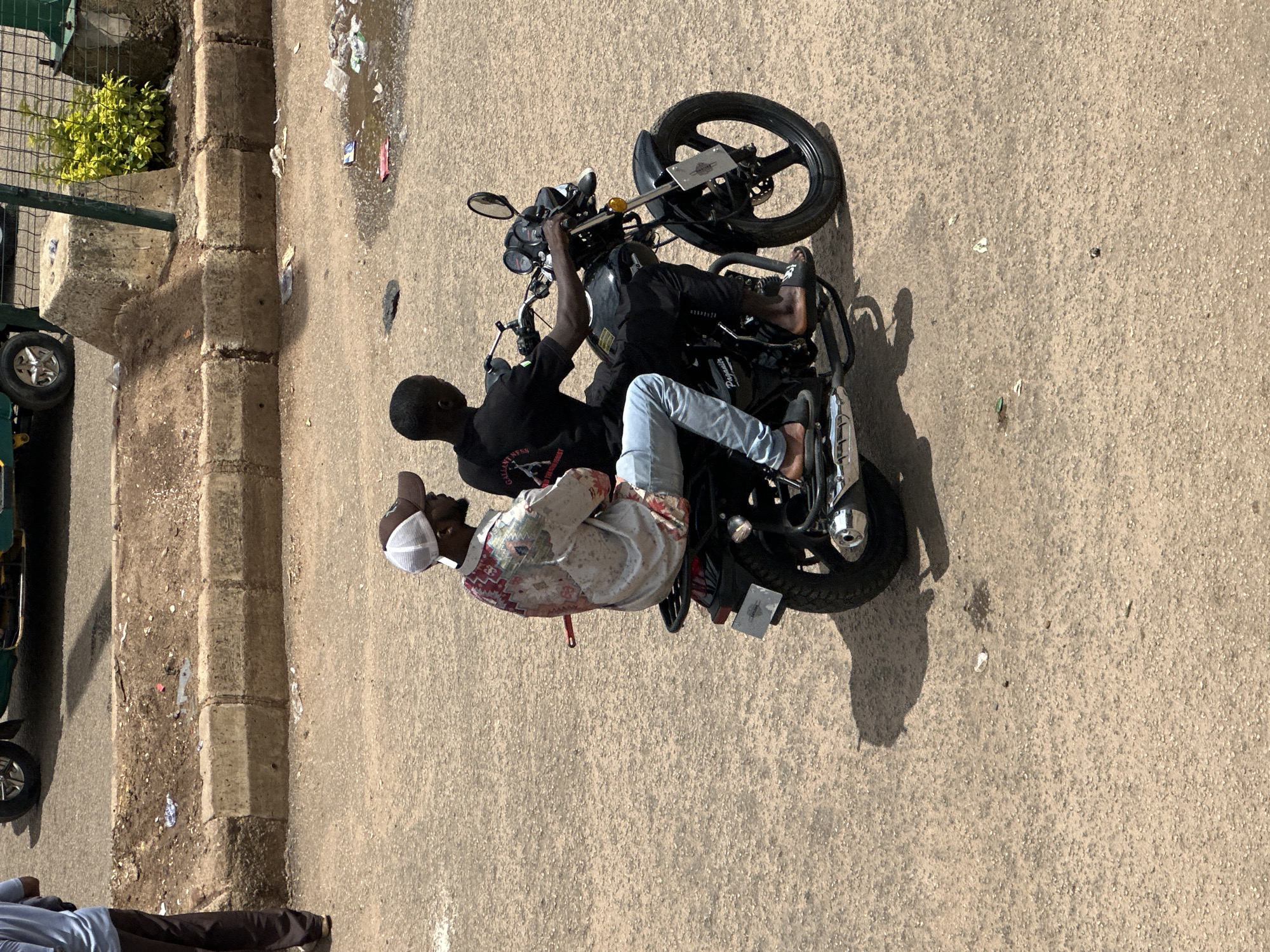 Two individuals are riding on a black motorcycle on a paved road. The rider in the front is wearing a black t-shirt and black pants, with a white logo on the back of his shirt. He is looking forward, operating the motorcycle. The passenger behind him is wearing a patterned shirt and light blue jeans. He is looking to his right. Both individuals appear to be adults. The motorcycle has a license plate that is partially visible. The road surface is rough asphalt. To the left of the road is a raised concrete curb with dirt and debris on top. Beyond the curb is a dirt area with some green shrubbery. In the background, a portion of a vehicle is visible, along with a chain-link fence. The scene appears to be an outdoor urban environment.