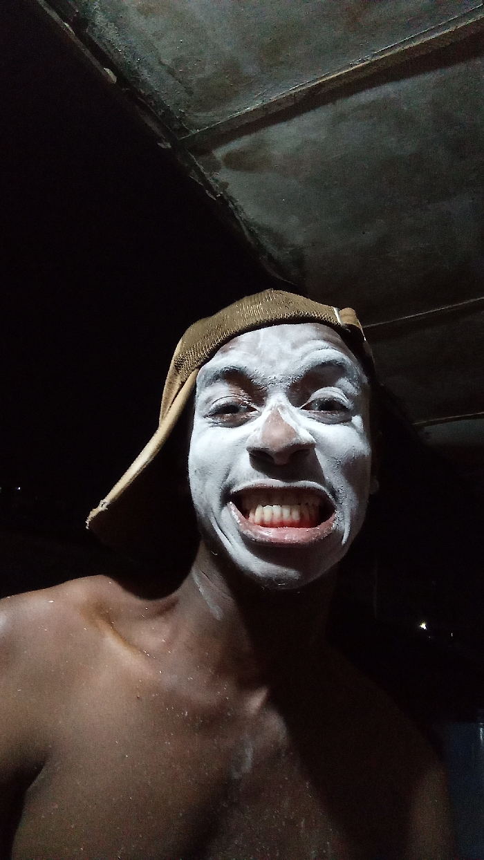 A dimly lit portrait features a young individual with dark skin, captured in a close-up, head-and-shoulders shot. The person is looking directly at the camera with wide, expressive eyes and a broad, toothy grin, revealing both upper and lower teeth. Their face is covered in a thick, white substance, possibly paint or a mask, which starkly contrasts with their natural skin tone and highlights their features. A brown, textured cap is worn on their head. The individual appears shirtless, with their shoulders and upper chest visible, showing a light sheen on the skin, possibly from moisture or the applied substance.
The background is predominantly dark, suggesting either a nighttime setting or a poorly lit indoor space. Above the person's head, a dark, textured surface, possibly a ceiling or roof, is partially visible, indicating a sheltered environment. The artificial lighting, likely a flash, illuminates the subject's face prominently. The person's exaggerated smile conveys a sense of cheerfulness, excitement, or playfulness. This image, possibly taken in Onitsha, Nigeria, could depict participation in a local event, a costume, or a moment of whimsical self-expression. No text is visible in the media.