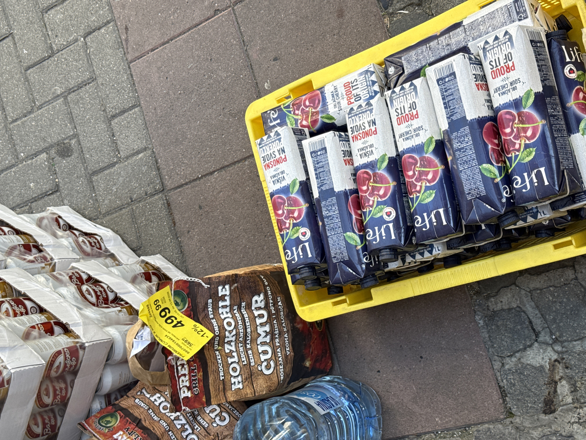 The image shows a collection of goods on a paved surface, likely a sidewalk or similar area in Belgrade, Serbia. The items include several packages of "Life" brand sour cherry juice in a yellow crate, bags of "Premium Cumur" grill charcoal with a price tag of 499.99 RSD, cases of beer cans, and a bottle of water. The arrangement suggests a recent purchase or delivery, with items scattered on the ground.