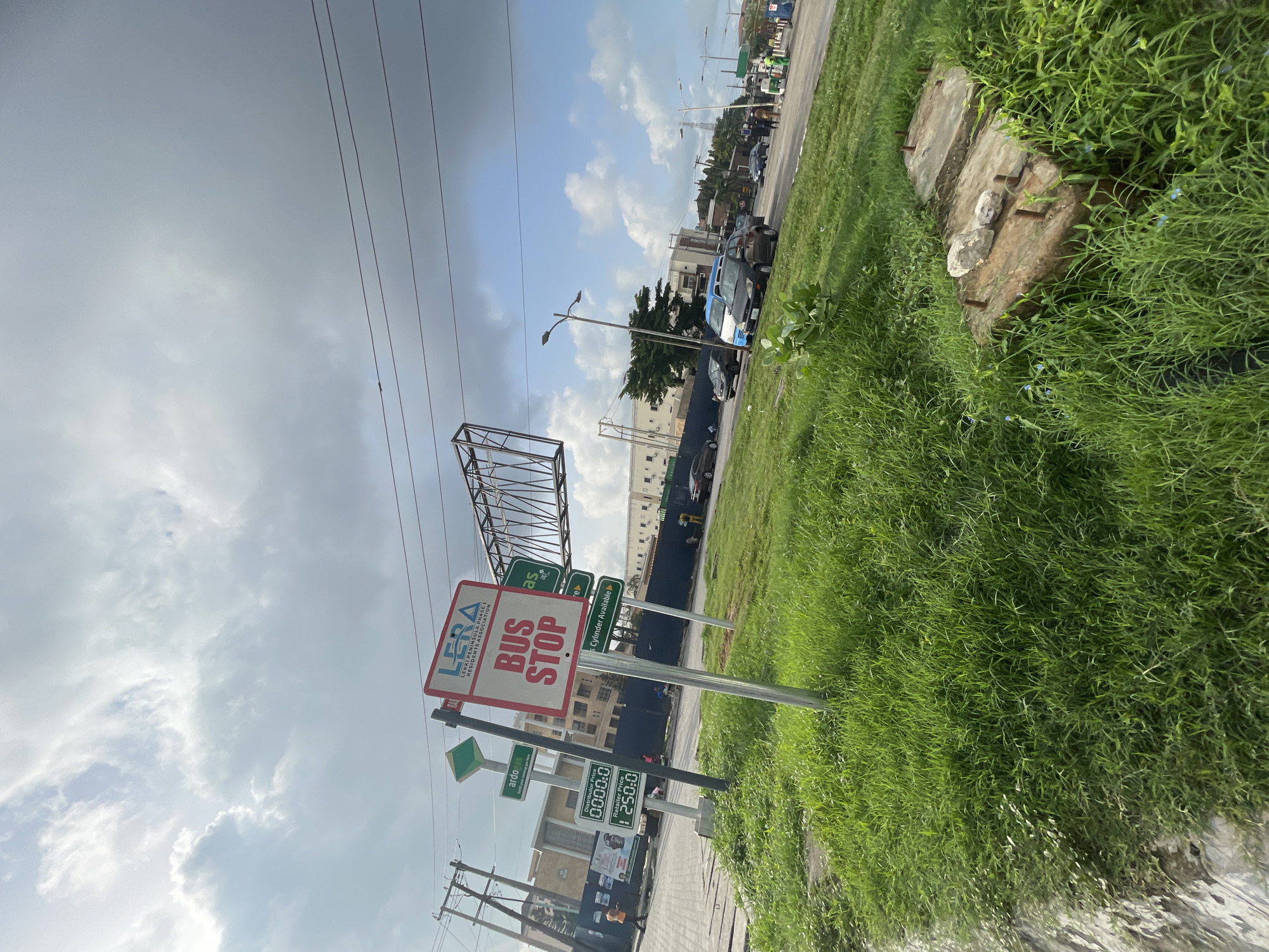 The image shows a bus stop sign in Lekki Peninsula Phase 1, Moba, Nigeria. The sign has the logo of LERA (Lekki Peninsula Residents Association) and states "BUS STOP" in large letters. Below the bus stop sign are signs indicating prices for distributors and retailers, displaying "0000:0" and "250:0", respectively. There are also other signs with text that is partially obstructed. In the background, there is a street with cars parked along the roadside. Lush green grass borders the street. There are utility poles with power lines visible in the sky, which is partly cloudy. There's a partially constructed or disassembled metal structure with visible crossbeams.