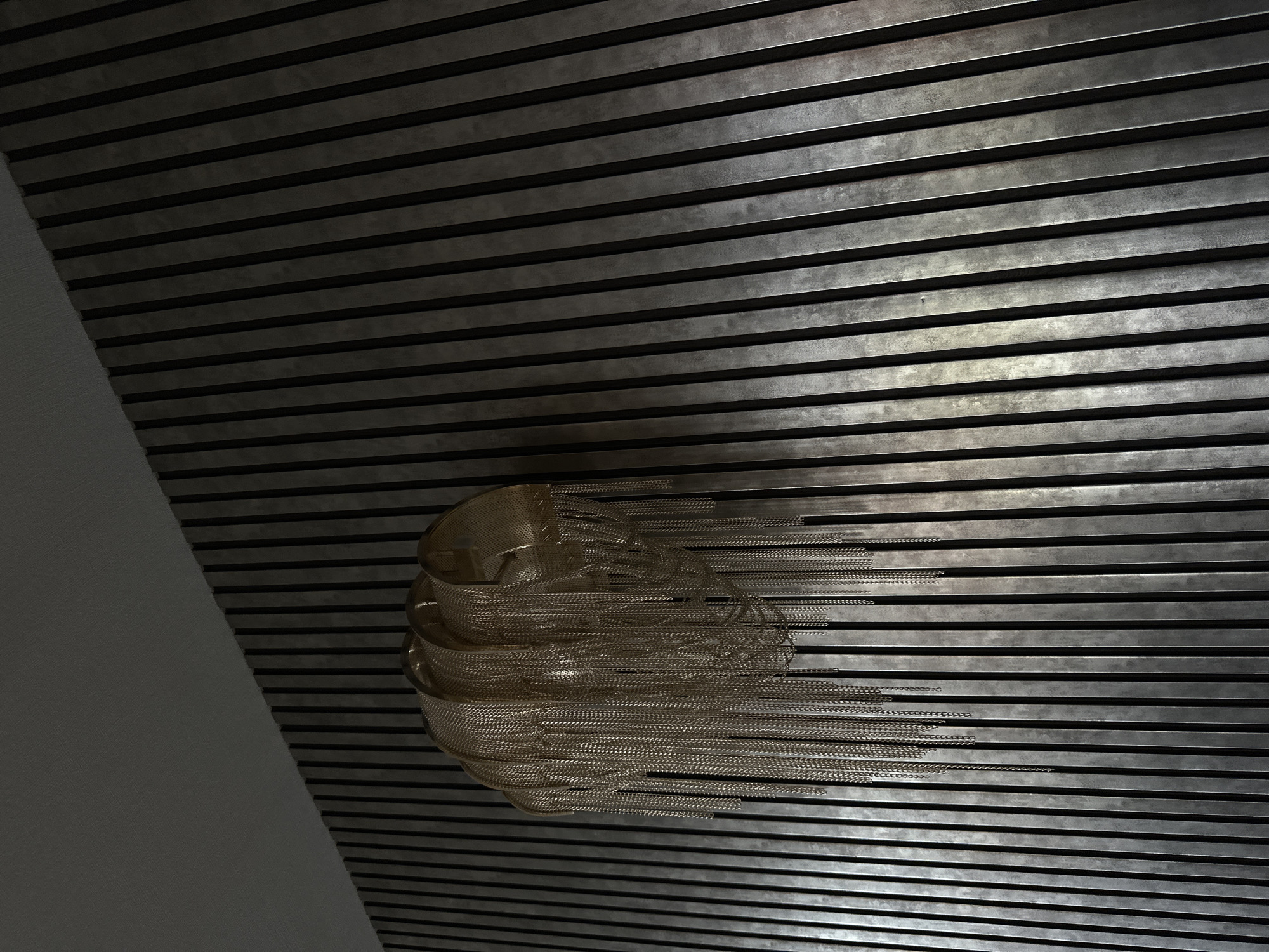 An eye-level, medium shot shows an interior wall with horizontal metal slats. The wall is dark and has a metallic sheen. On the left side of the wall, an adjacent white wall is visible at an angle. A sculptural, metallic, sphere-like object with trailing chain-like strands is attached to the slatted wall. The photo was taken indoors, potentially in Marneuli, Georgia.