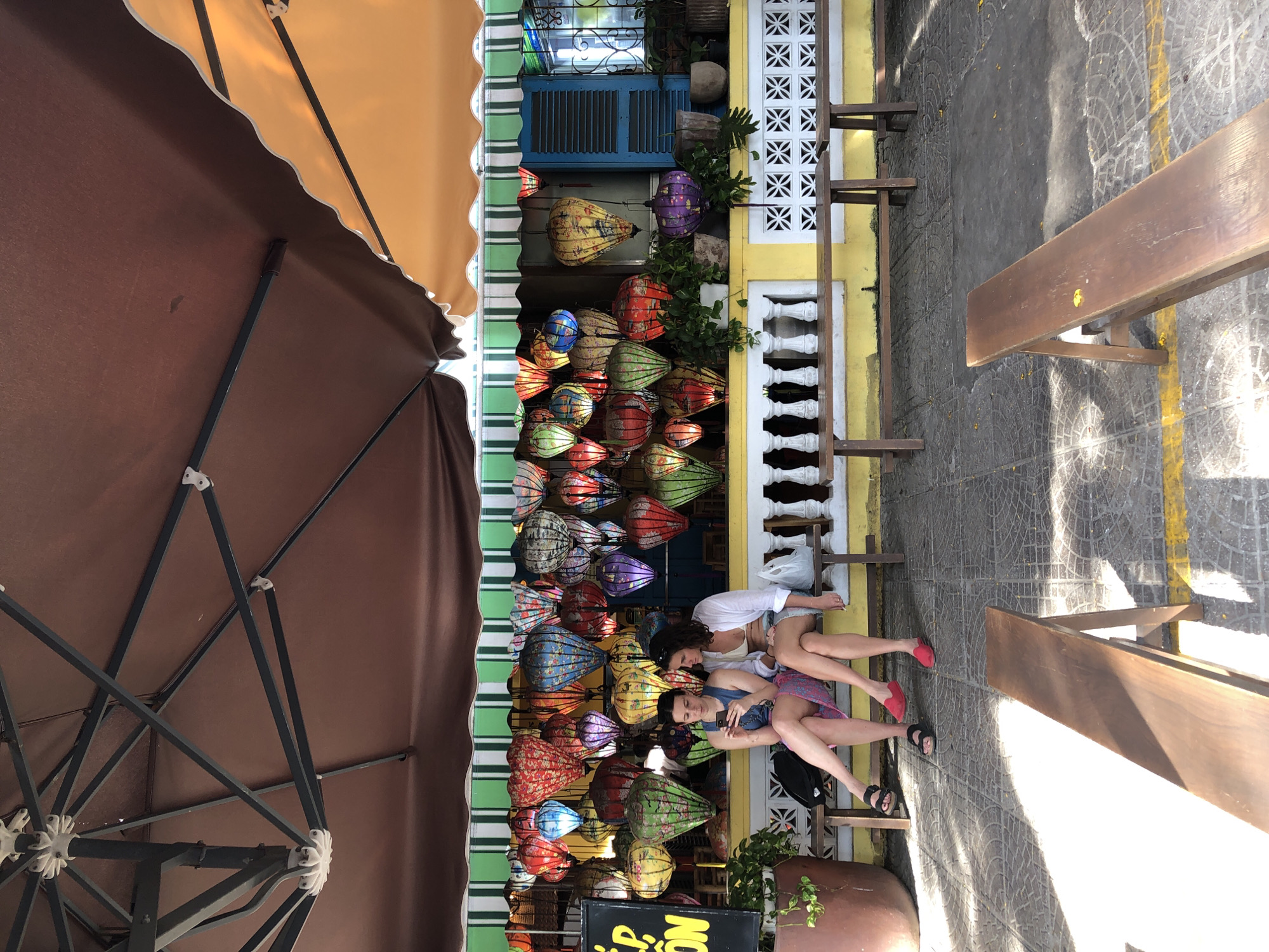In Đà Nẵng, Vietnam, two women are sitting on a wooden bench under a brown awning. The woman on the left is wearing a blue sleeveless top and a patterned skirt, while the woman on the right is wearing a white open shirt over a bikini top and denim shorts. They are both looking at a smartphone. Behind them, a wall is decorated with numerous colorful, illuminated lanterns. The wall is painted yellow, and there's a green and white striped awning above it. To the left of the lanterns, a large brown planter holds a green plant. In front of the bench, two more wooden benches are visible. The ground is paved with patterned tiles and includes yellow line markings. The scene appears to be an outdoor seating area in front of a shop.