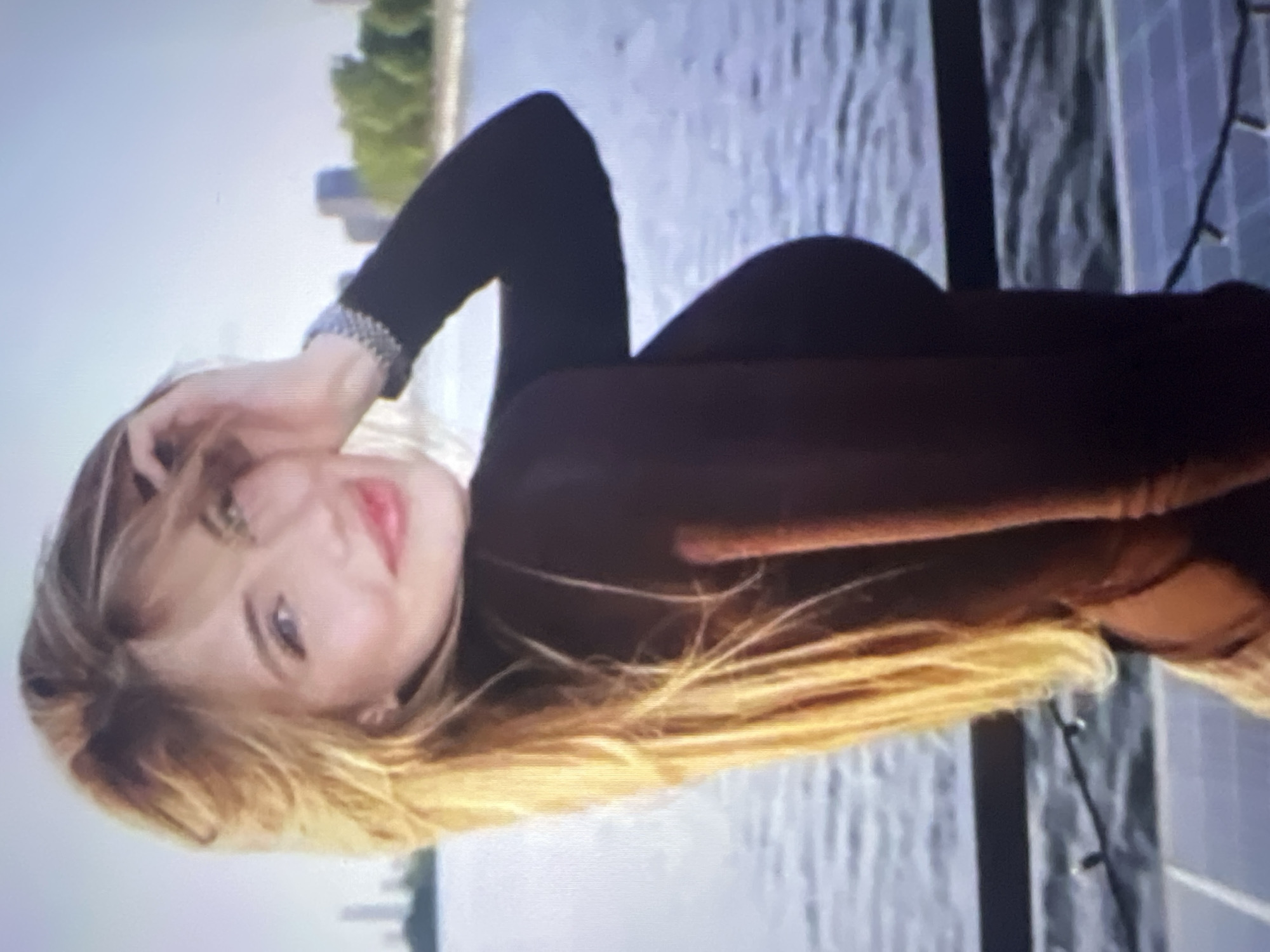 A close-up shot shows a young woman with long blonde hair styled in layers, her right hand touching her hair. She is wearing a dark, long-sleeved top. Her left arm is bent, with her hand resting on her hip. She is looking towards the camera with a soft smile and her lips slightly parted. In the background, a body of water stretches out, with a faint skyline of buildings and green trees on the horizon. The light suggests it's daytime, likely late afternoon or early evening, with the sun casting a warm glow. The overall atmosphere is relaxed and casual. There is no indication of Energetik, Russia in the image.