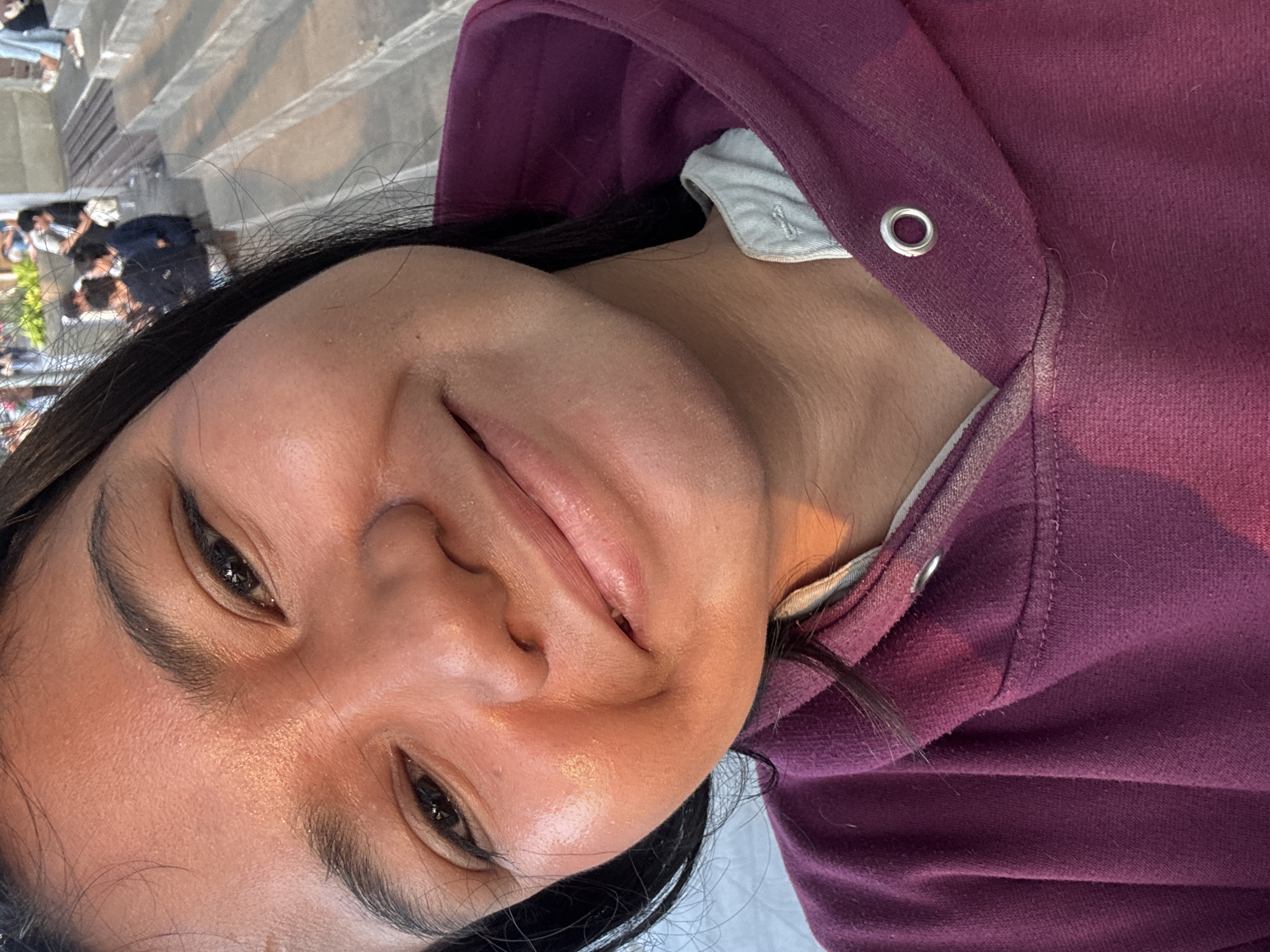 A close-up selfie shows a young woman with dark hair smiling at the camera. She is wearing a deep purple hooded sweatshirt with a light grey collar. The background is blurred, but appears to be an outdoor public space with tiered seating, possibly stairs or bleachers. In the distance, several people are visible, some sitting and others standing. The lighting suggests it is late afternoon or early evening, with the sun casting a warm glow on the woman's face. The setting is likely Ancol Beach City, North Jakarta, Indonesia, a popular recreational area with boardwalks and entertainment venues.