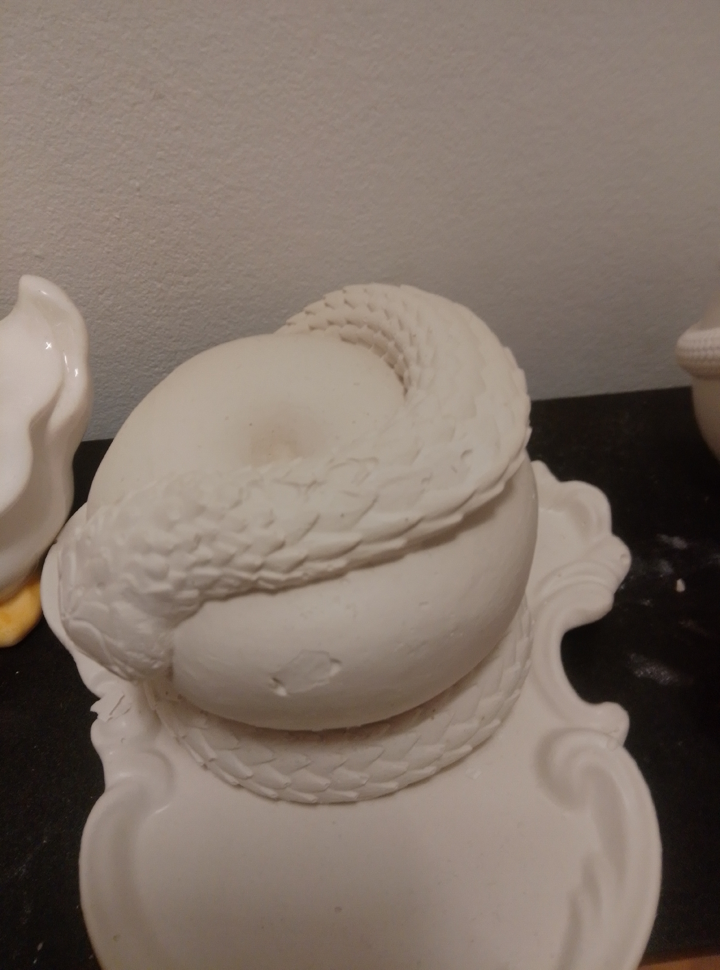 The image shows a white ceramic sculpture of a sphere wrapped in a coiled snake, resting on an ornate white ceramic base. The sculpture is unpainted and appears to be unfinished. To the left of the sculpture, a portion of another white ceramic object with a yellow base is visible. The objects are placed on a dark surface, possibly a table, in front of a textured, light-colored wall. The scene is indoors and appears to be in a domestic setting. The location of Wedel, Germany is not depicted in the image.
