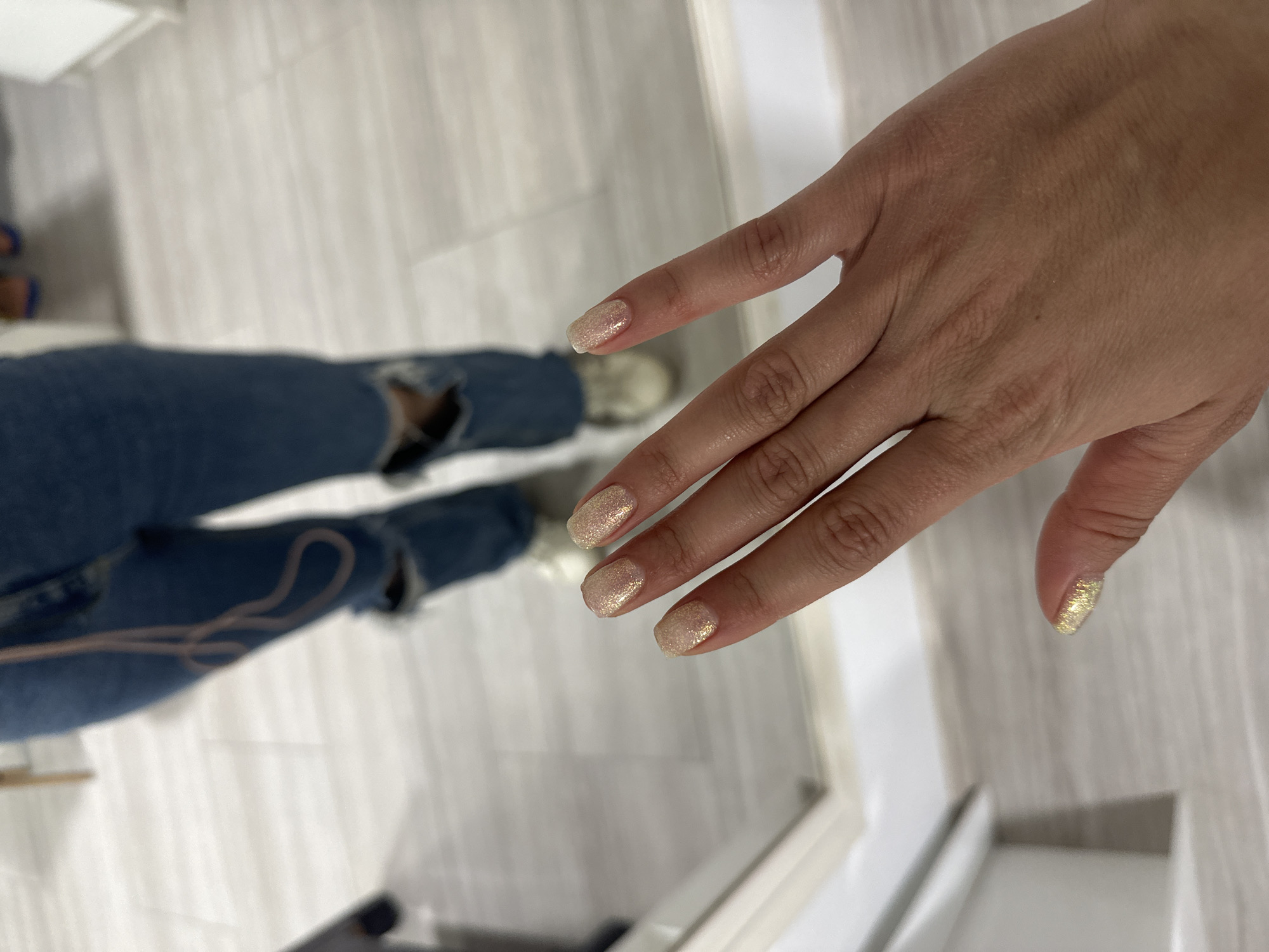 This media file shows a close-up view of a person's hand with glittery, light-colored nail polish. The hand is positioned in the foreground, with the fingers slightly curled. The background is out of focus, but appears to be an indoor setting with a light-colored, possibly tiled floor.
In the middle ground, a person's legs are visible, wearing ripped blue jeans. A thin, light-colored string or strap is draped across the jeans. The person is wearing sneakers.
The lighting suggests it is daytime. The overall scene appears casual, possibly taken in a home or a salon. There is no visible text in the image. The location context provided is Haskovo, Bulgaria.
