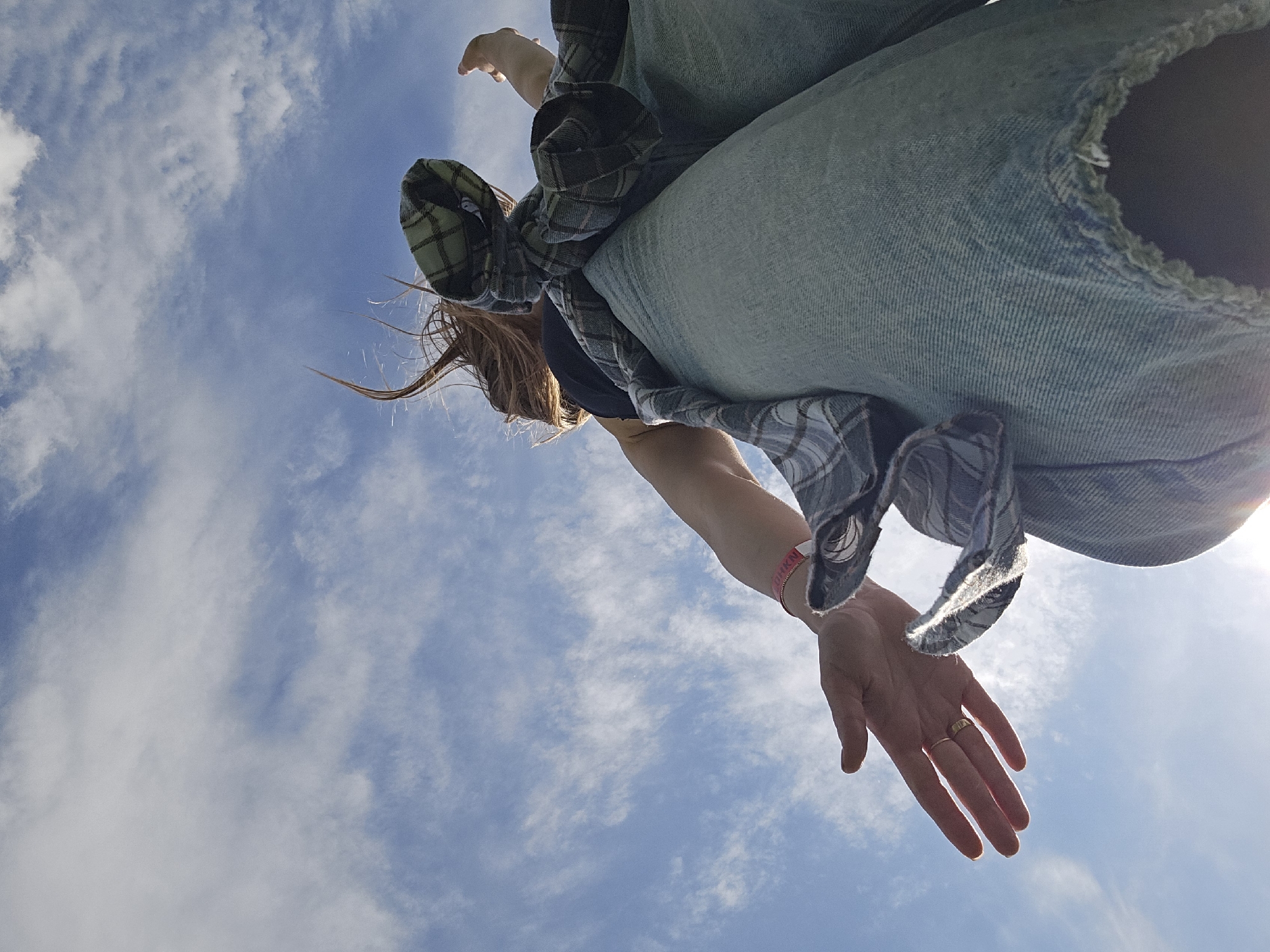 The image shows a low-angle shot of a person against a blue sky with scattered clouds. The person is wearing ripped light blue jeans, a black t-shirt, and a green and grey plaid shirt tied around their waist. The person's left arm is visible, extending towards the camera, with a pink wristband and a gold ring on the ring finger. Strands of brown hair are also visible. The sun is bright in the lower right corner. The location is not identifiable as Izhevsk, Russia based solely on the visual information in the provided image.
