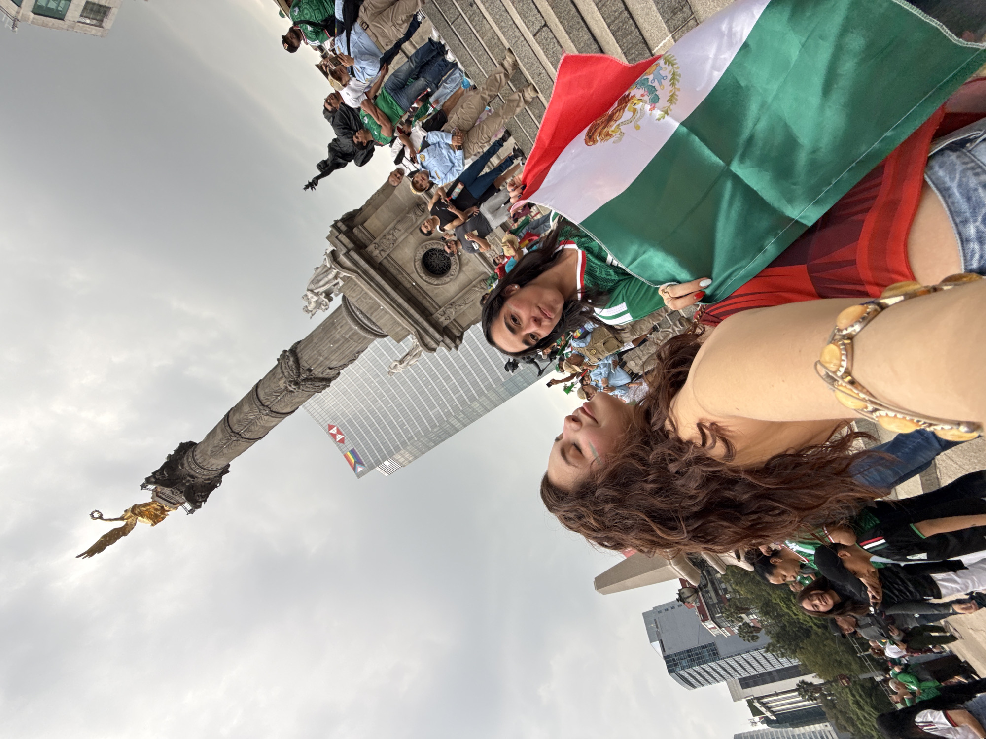The image depicts a group of people gathered at the Monumento a la Independencia in Mexico City. In the foreground, two women are prominent. One woman, with long, curly brown hair, is holding a bracelet with yellow beads and has paint, possibly face paint, on her cheek. The other woman is holding a Mexican flag, which is predominantly green, white, and red, with the Mexican coat of arms visible. She is wearing a green and white jersey.
In the background, the Monumento a la Independencia, also known as El Ángel, stands tall with its golden sculpture of the Winged Victory of Samothrace at the top. The monument is made of stone and features intricate carvings and statues at its base. Several other people are visible, some wearing green jerseys, suggesting a gathering related to a sporting event or national celebration. Behind the monument, modern buildings are visible, including a skyscraper with a logo that appears to be for HSBC. The sky is overcast with clouds. The scene appears to be an outdoor public gathering in a city square.