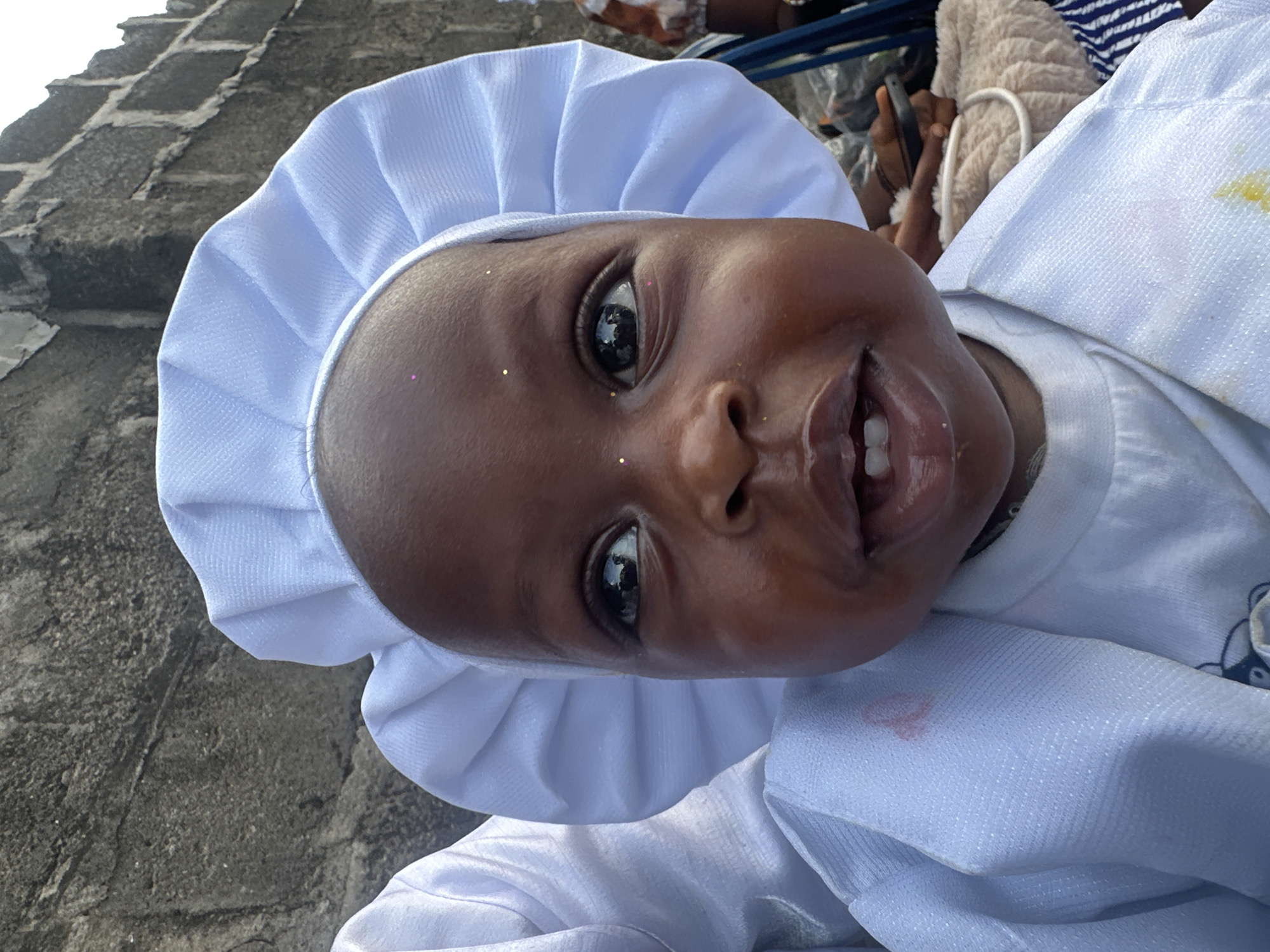 The image captures a young child with dark skin, positioned in the foreground and looking directly at the camera with wide-open, dark eyes. The child wears a white, pleated or ruffled bonnet and a white long-sleeved outer garment, which appears to be layered over another white top. Small, reflective, glitter-like specks are visible on the child's forehead and left cheek. A light reddish-pink stain is present on the upper left shoulder area of the outer white garment, while a yellowish stain is seen on the lower right front of the same garment. The child's mouth is slightly open, revealing two lower and two upper front teeth.
The background consists of a rough, grey wall constructed from what appears to be concrete blocks or large stones, showing distinct textures and mortar lines, particularly prominent behind the child's head and to the upper right. The top edge of this wall is jagged. To the immediate left of the child, another person's white garment is partially visible, suggesting close proximity. To the right of the child, blurred elements indicate the presence of other individuals and objects: a darker-skinned hand holding a black smartphone, a beige, fuzzy object, and blue and white striped fabric. The scene is situated in Badagry, Nigeria.