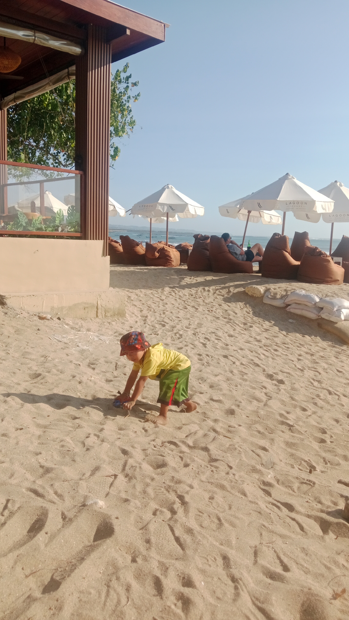 A young boy wearing a yellow t-shirt, green shorts, and a colorful patterned hat is playing on a sandy beach in Kuta, Indonesia. He is on his hands and knees, interacting with a toy. The beach is lined with white umbrellas and large brown beanbag chairs, some of which are occupied by other beachgoers. In the background, the ocean is visible under a clear blue sky. A wooden structure with vertical slats and a thatched roof is situated on the left side of the image, suggesting it is part of a beach club or restaurant. The lighting indicates it is daytime, with shadows cast by the boy and the nearby structure.